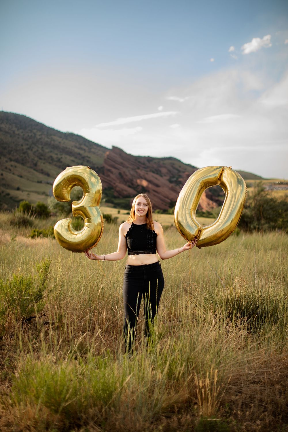 Smiling woman holding gold number 30 balloons in a grassy field with red rock hills, celebrating a 30th birthday photoshoot.