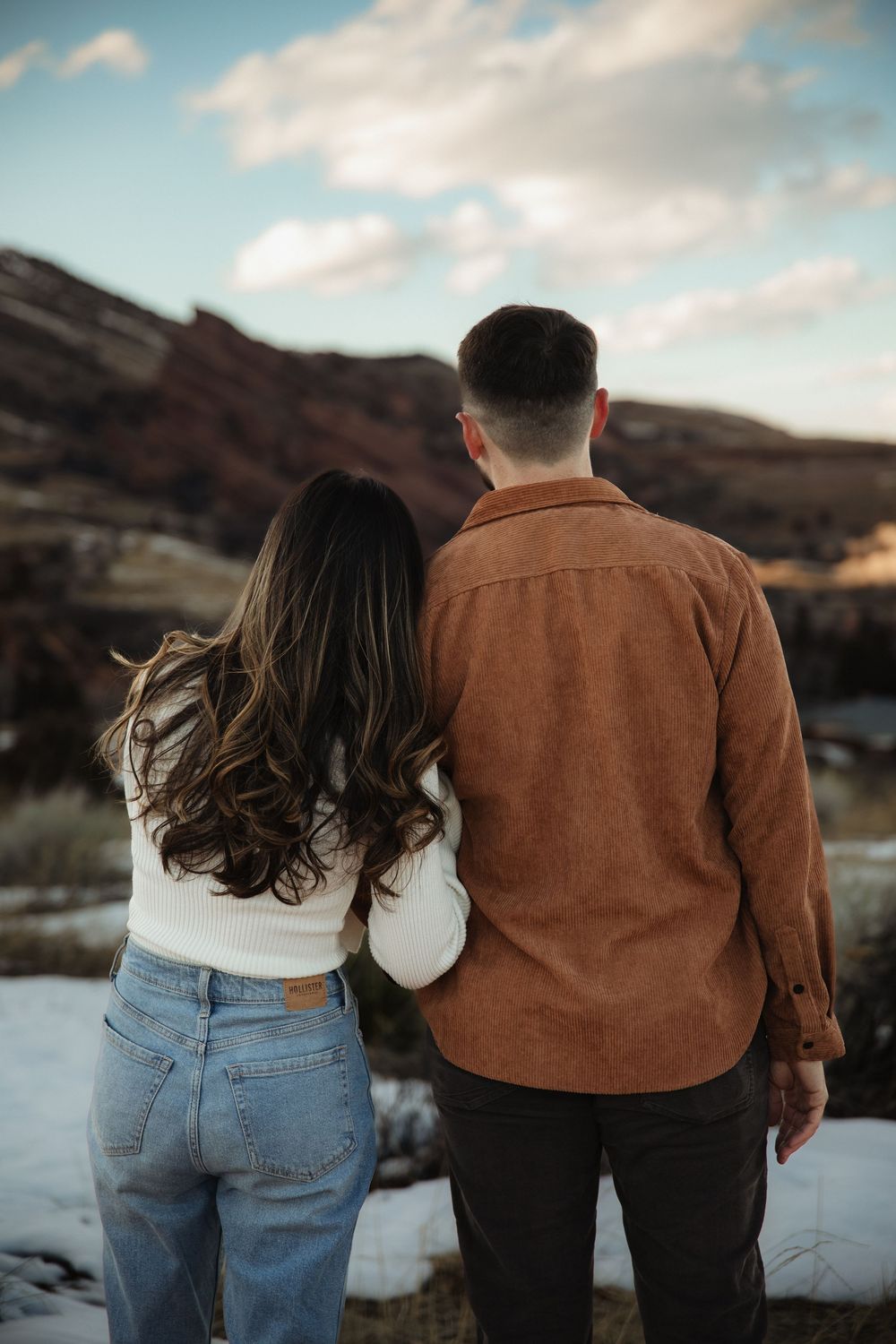Couple standing close with their backs to the camera, gazing at red rock cliffs and a snowy landscape under a cloudy sky.
