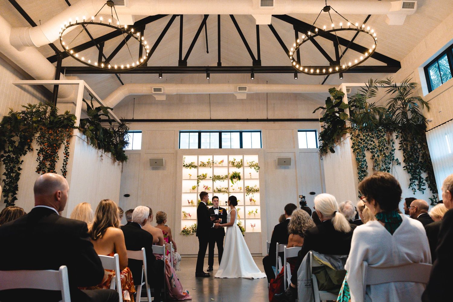 Joyful bridesmaids and groomsmen laugh and hug a bride holding a colorful bouquet inside a bright glass greenhouse wedding venue.