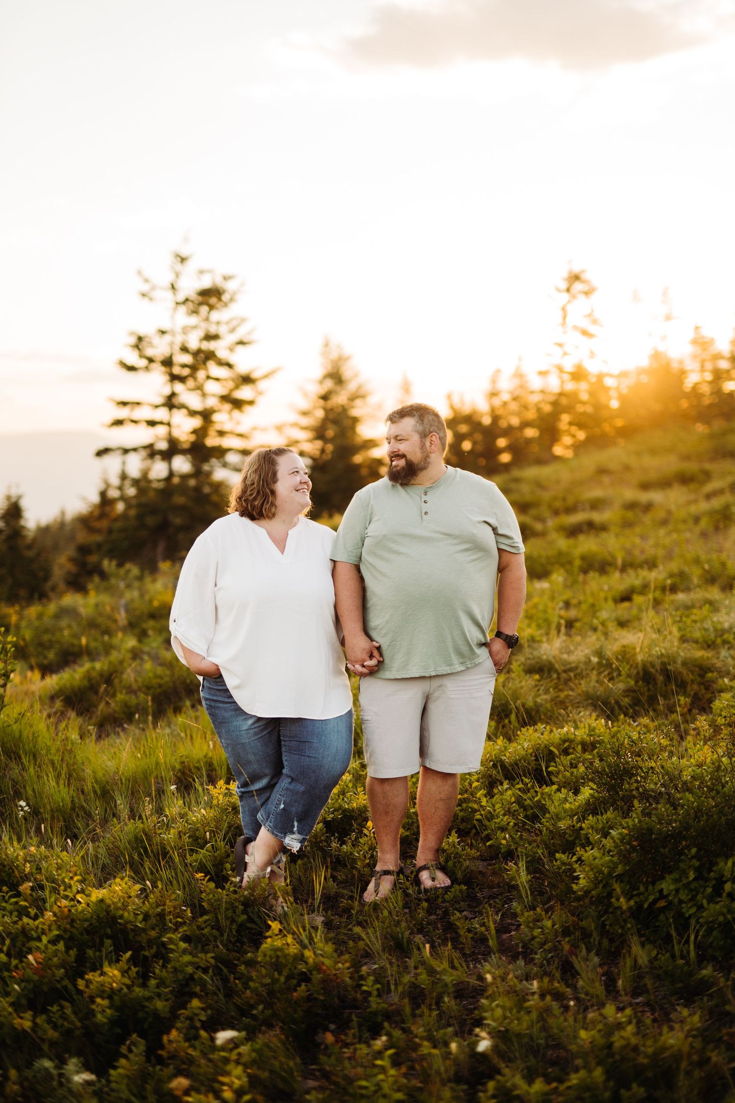 Couple holding hands smiling at each other in a sunlit meadow with pine trees at golden hour.