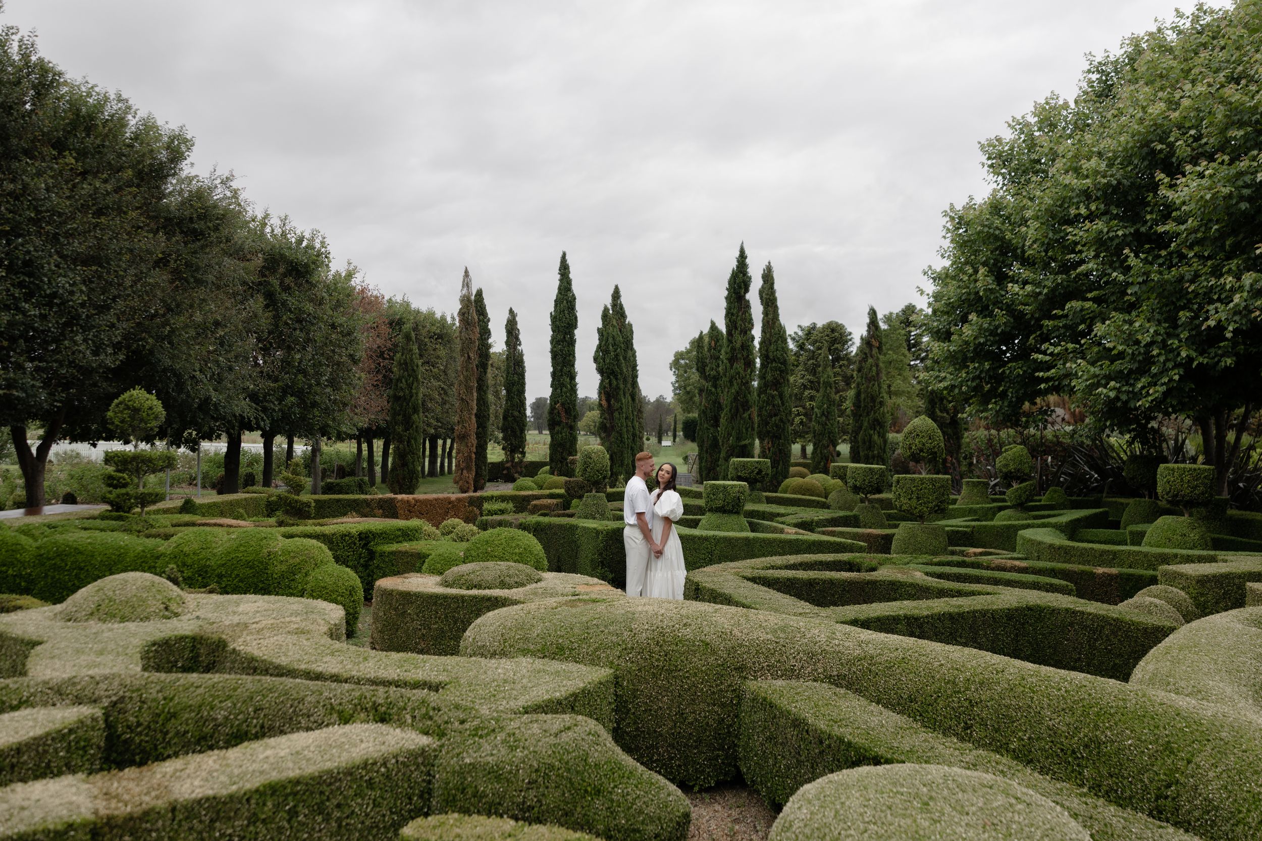 Couple embracing in a formal garden maze with sculpted hedges and cypress trees under an overcast sky.