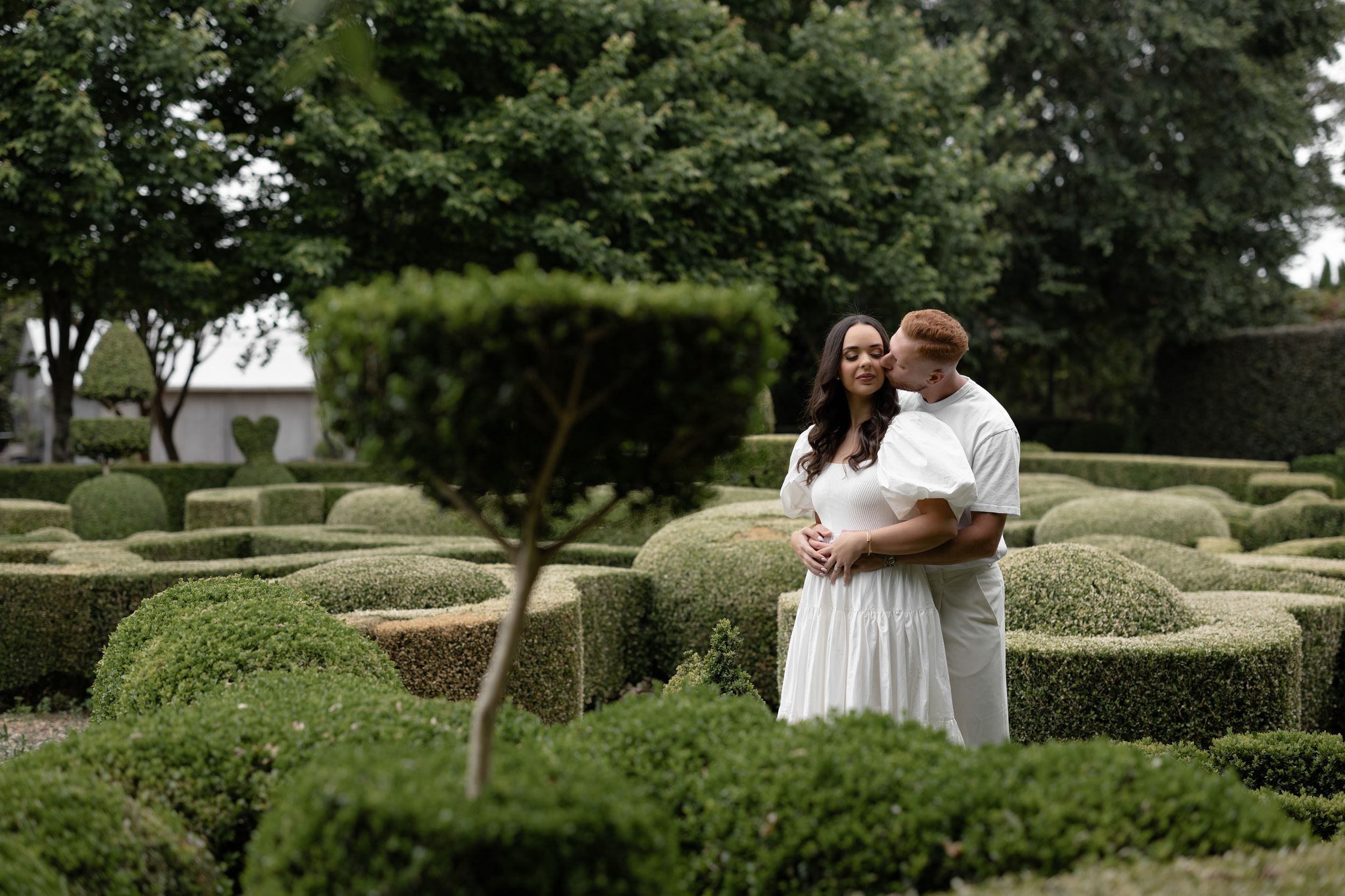 Couple embracing in a formal garden with neatly trimmed box hedges, woman in white dress, lush green surroundings.