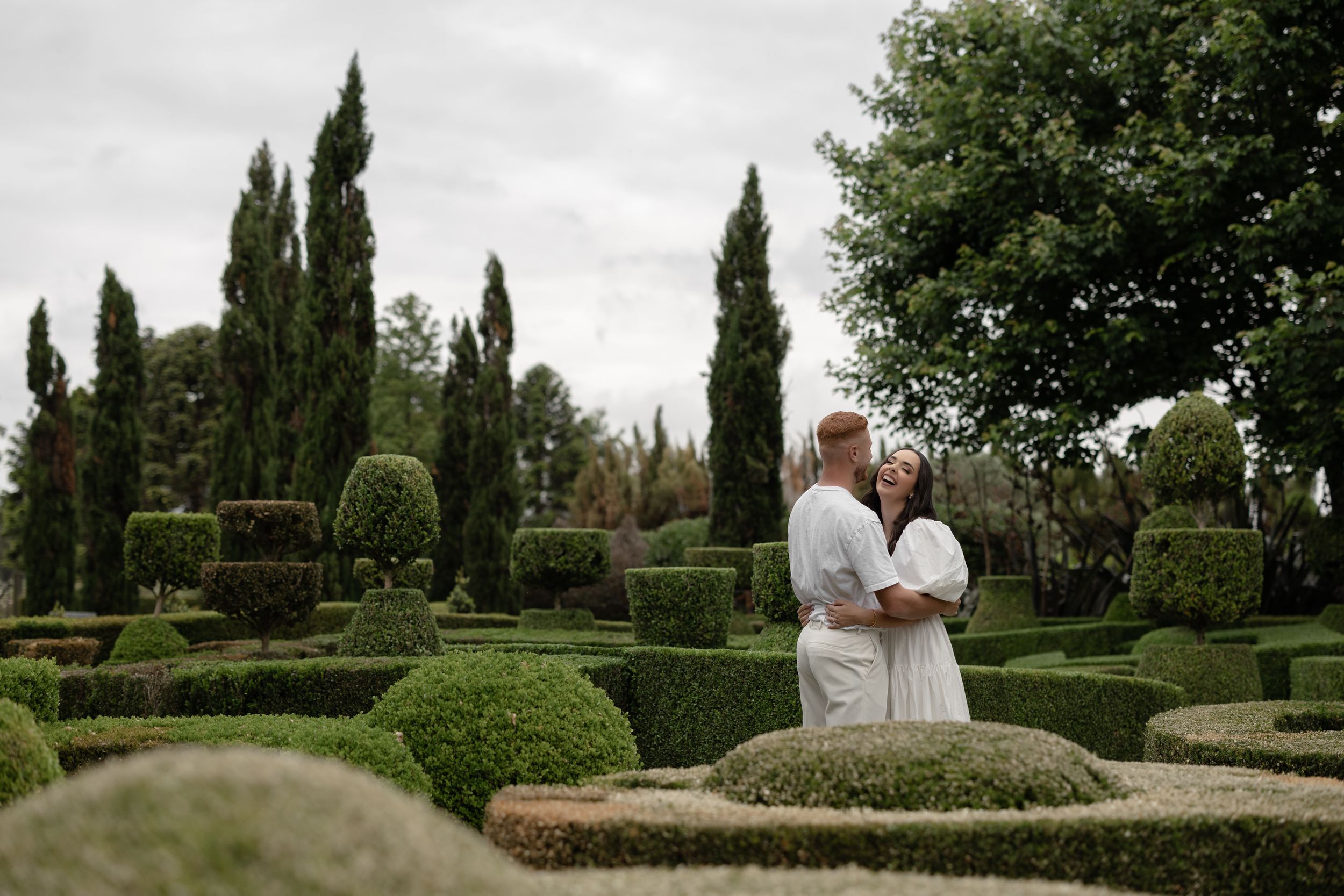 Couple embracing in a formal garden with manicured hedges and tall cypress trees on a cloudy day.