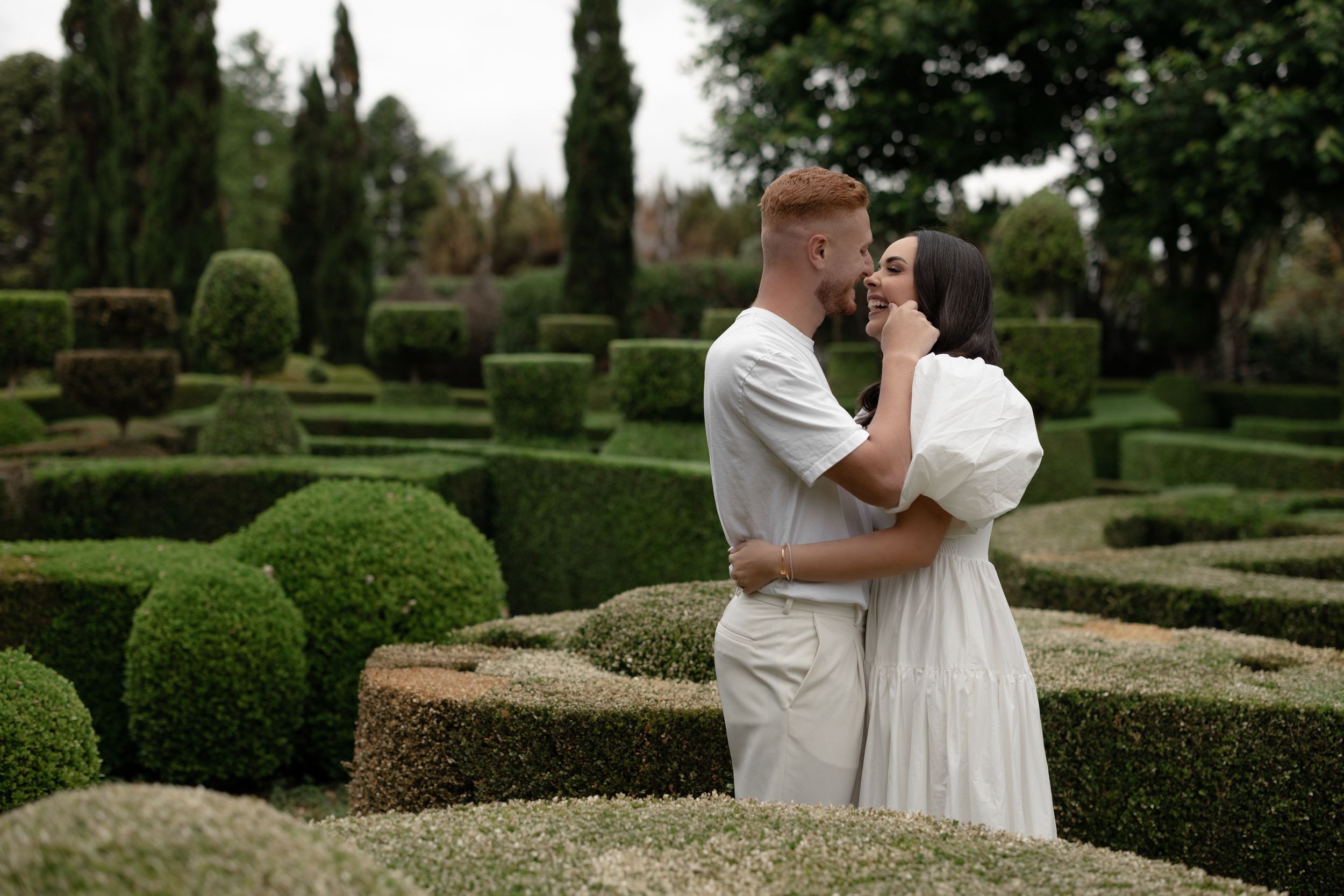 Romantic couple embracing and kissing in a lush formal garden with sculpted hedges and topiaries.