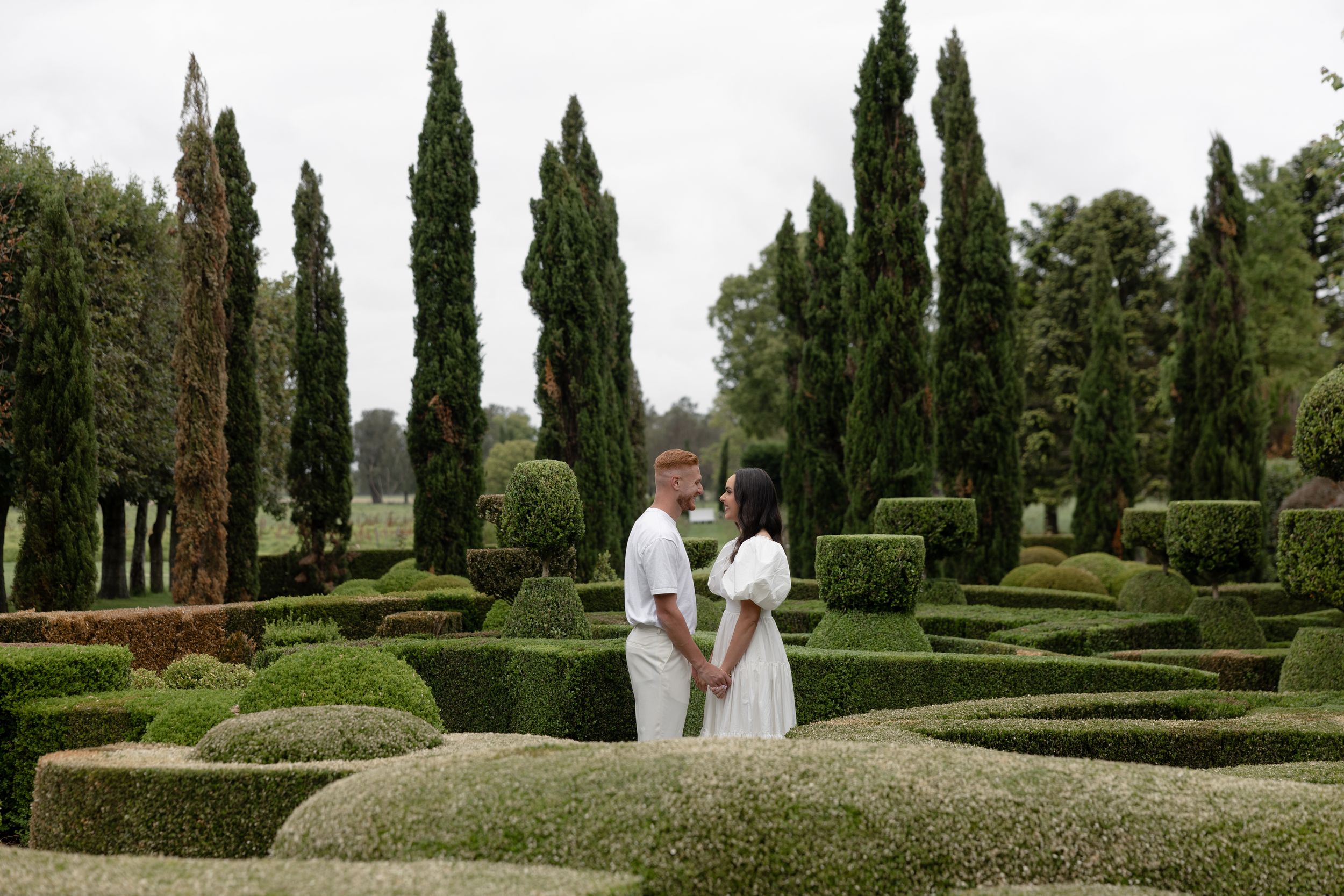 Couple in white attire holding hands in a formal garden with sculpted hedges and tall cypress trees.
