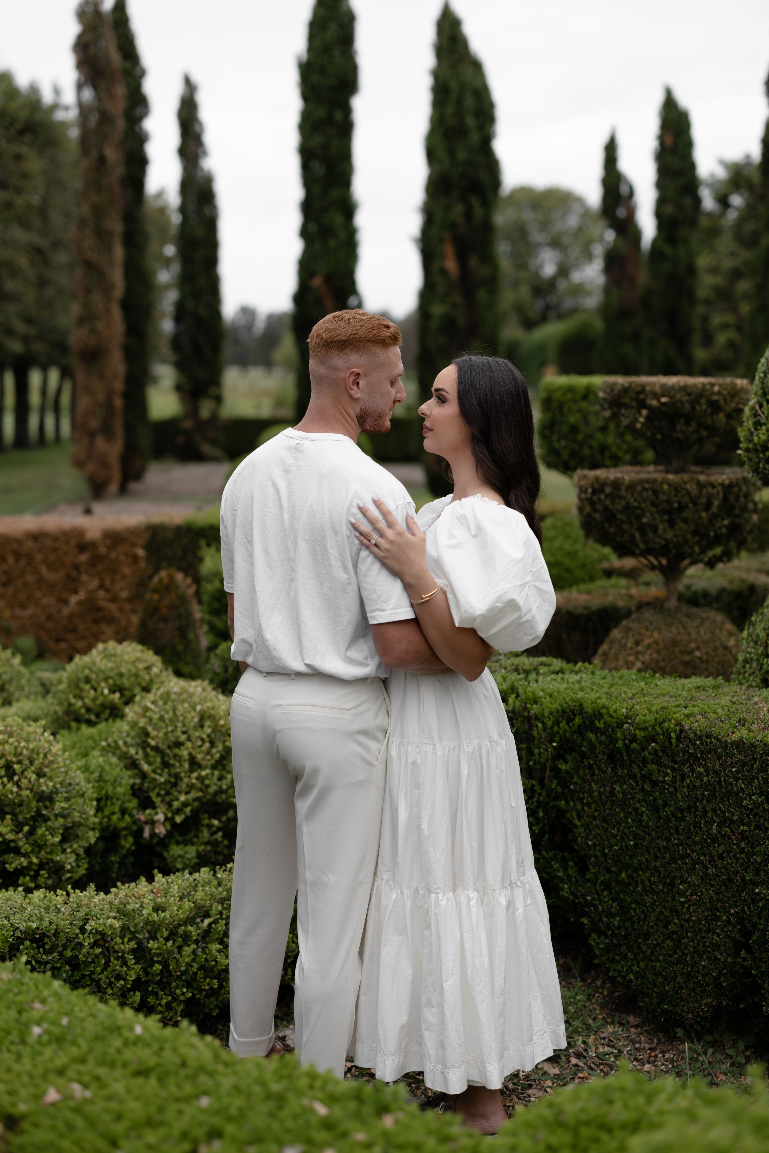 Couple in white outfits embracing in a formal garden with tall cypress trees and manicured hedges.