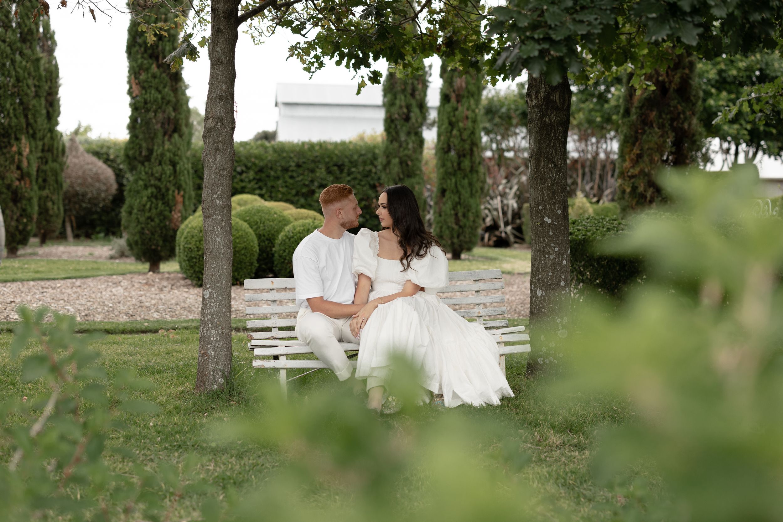 Couple in white outfits sitting on a white bench surrounded by lush green trees in a romantic garden setting.