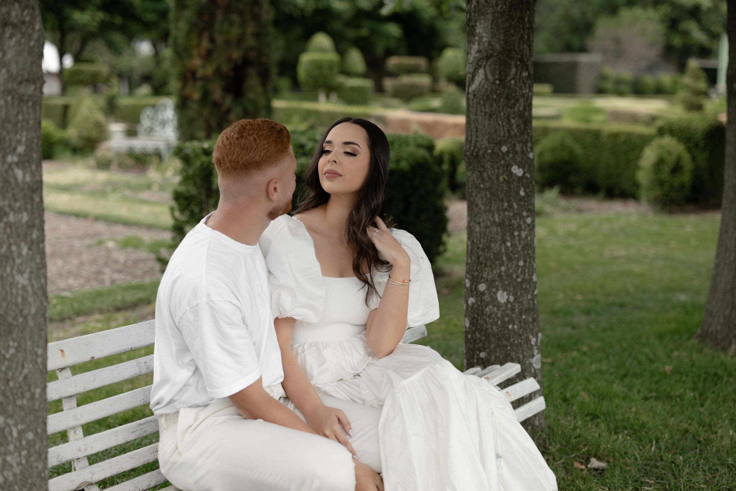 Couple in white outfits sitting on a bench in a lush green park, sharing a romantic moment.