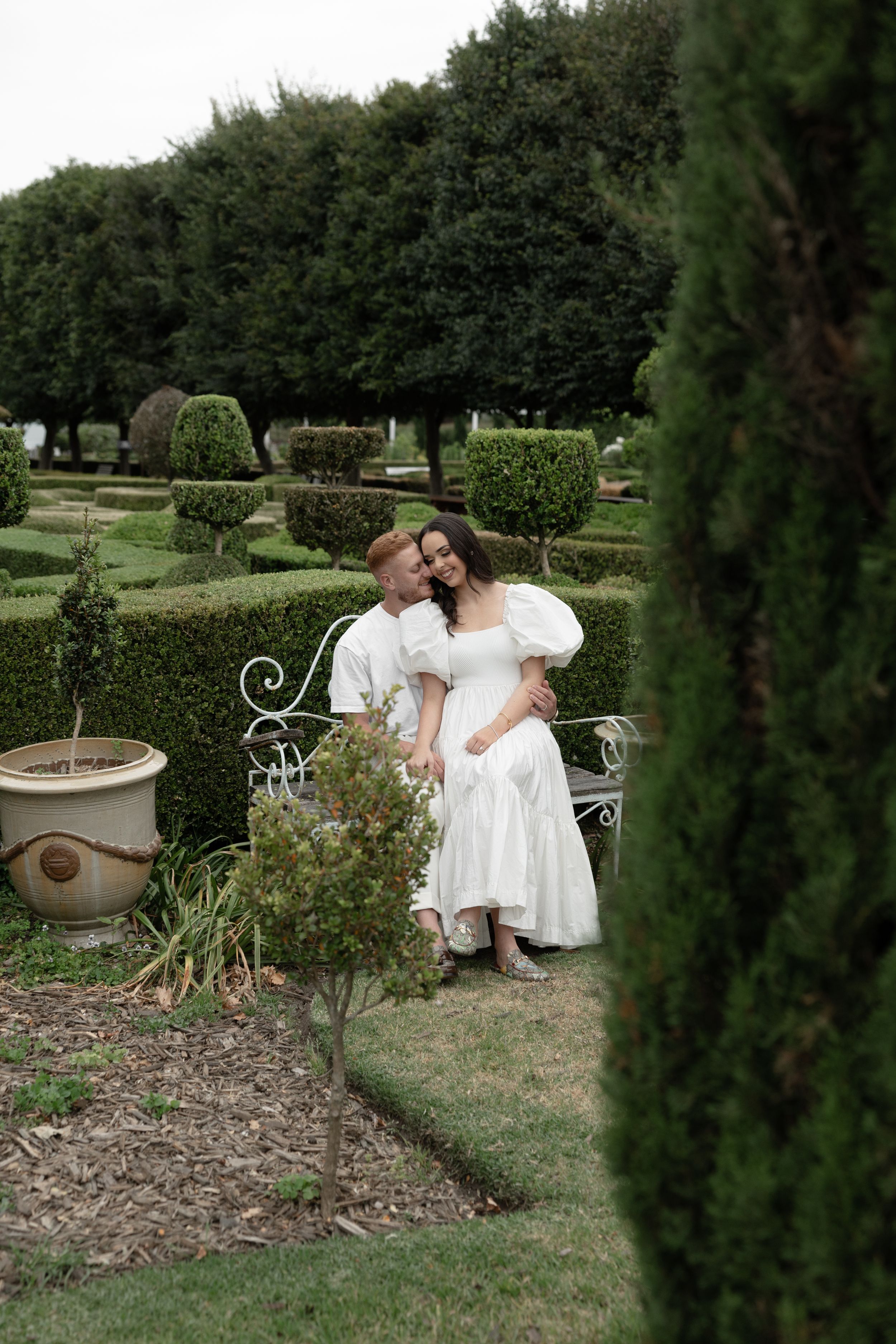 A couple in a formal garden, woman in white dress seated on ornate bench, man embracing her among manicured hedges.