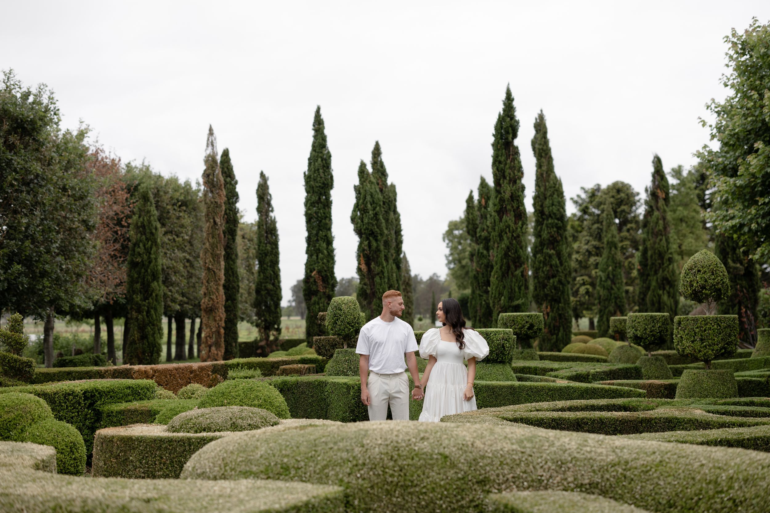 Couple in white outfits holding hands in a formal garden with manicured hedges and tall cypress trees.