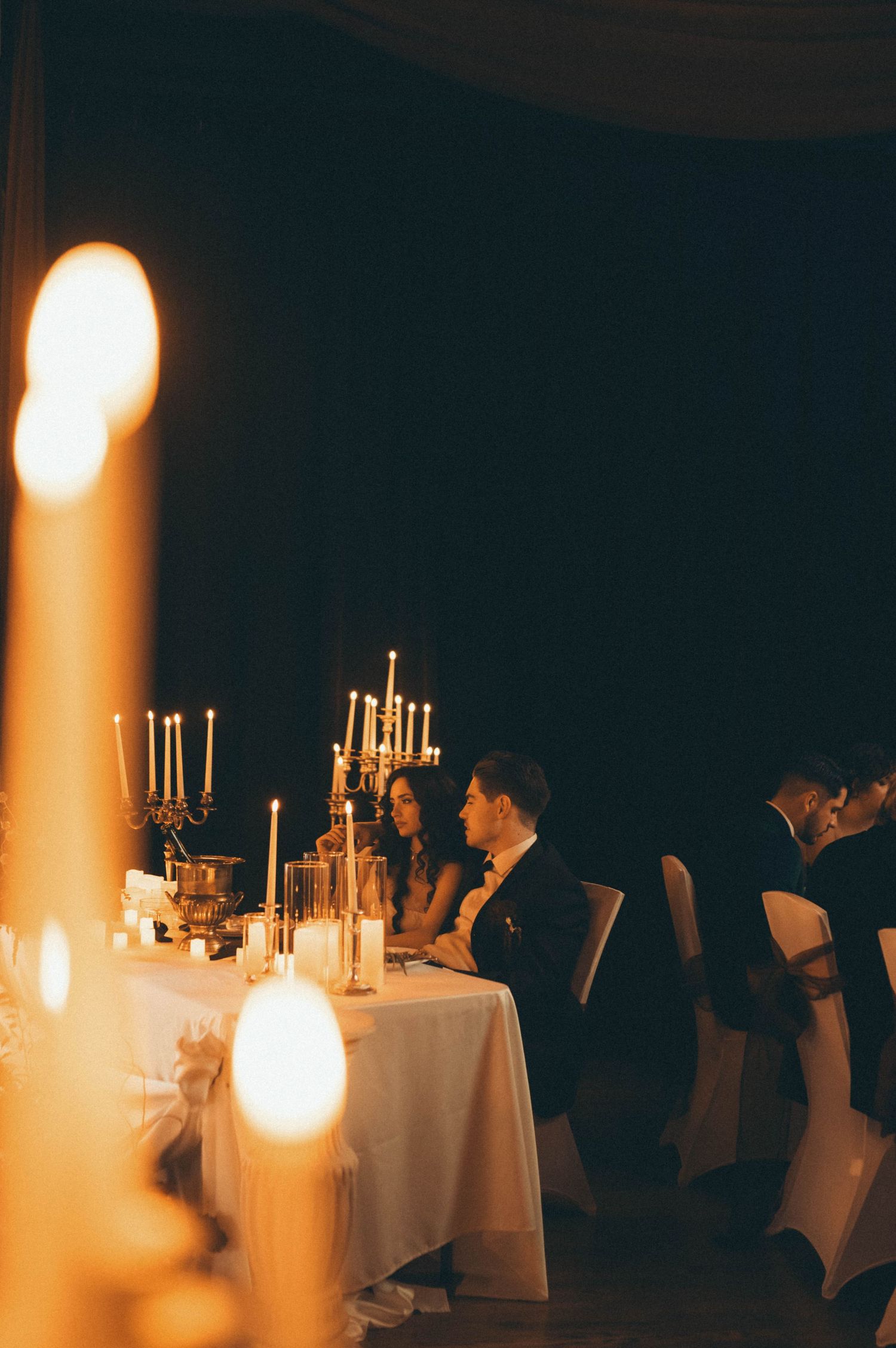 Couple seated at candlelit wedding reception table with elegant candelabras in a romantic, dimly lit venue.
