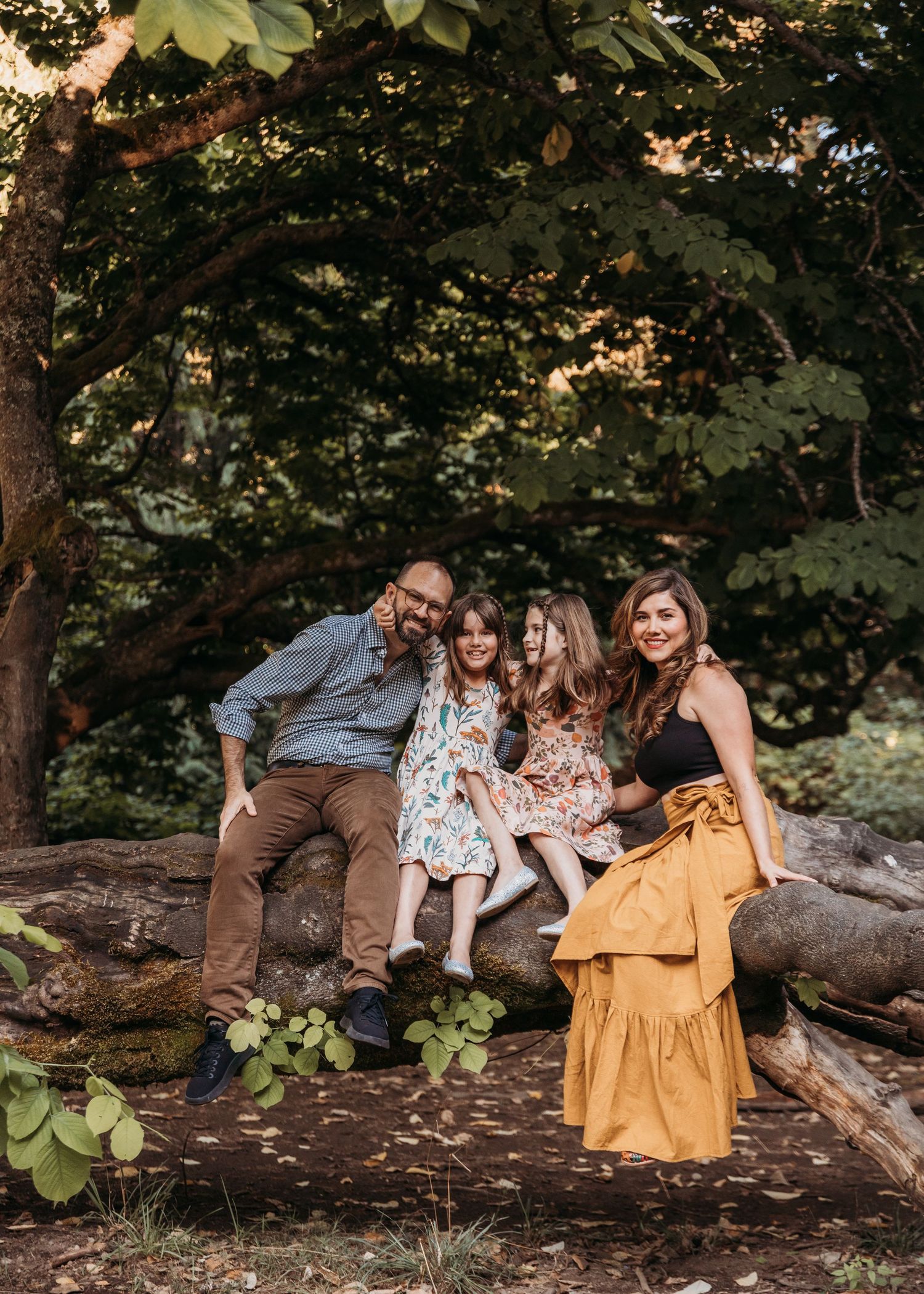Family of four sits together on a mossy fallen log in a sunny forest, with two young girls in floral dresses between their smiling parents.