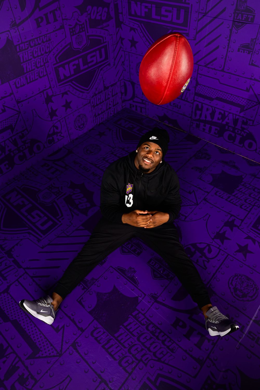 A smiling man in a black hoodie and beanie sits on a purple NFL-themed floor, looking up at a football tossed in the air.