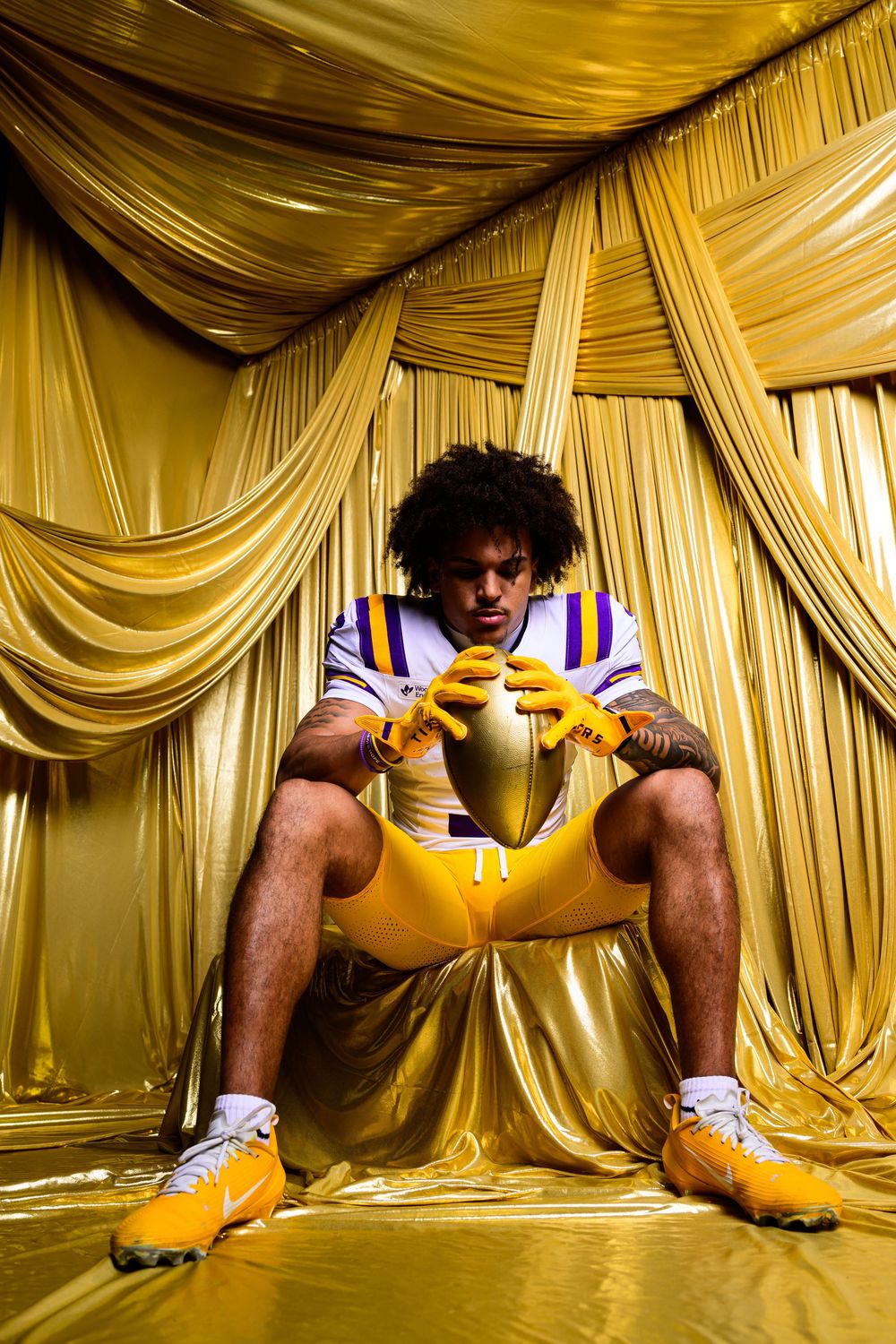Football player in gold uniform holds ball, seated against golden draped curtain backdrop.