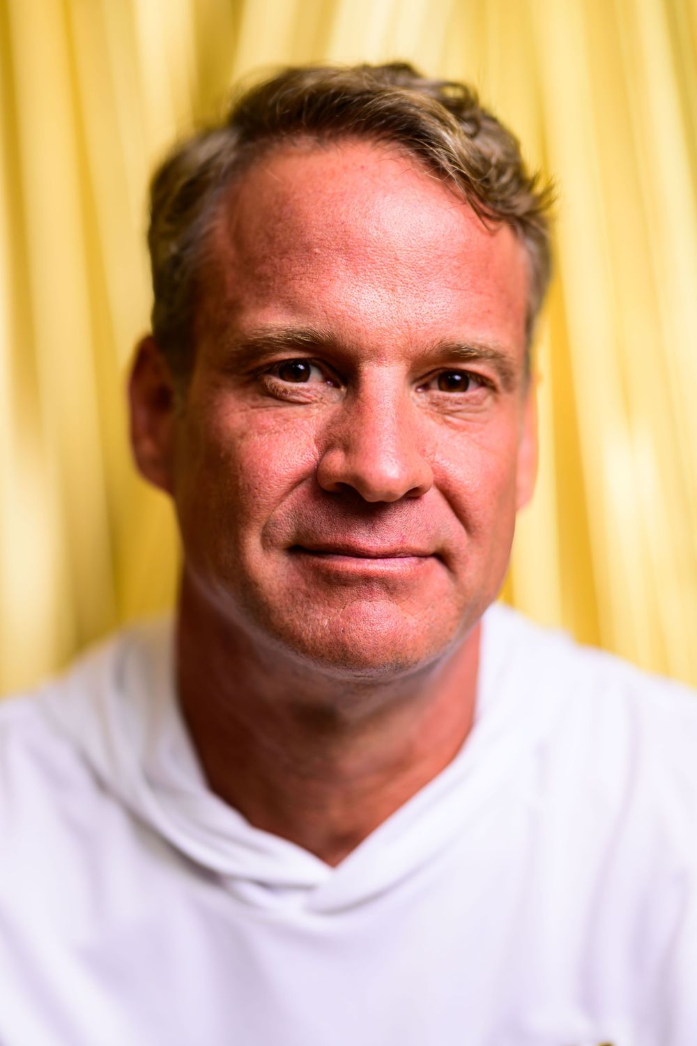 Smiling middle-aged man with short hair wearing a white hoodie, posed against a yellow curtain background.