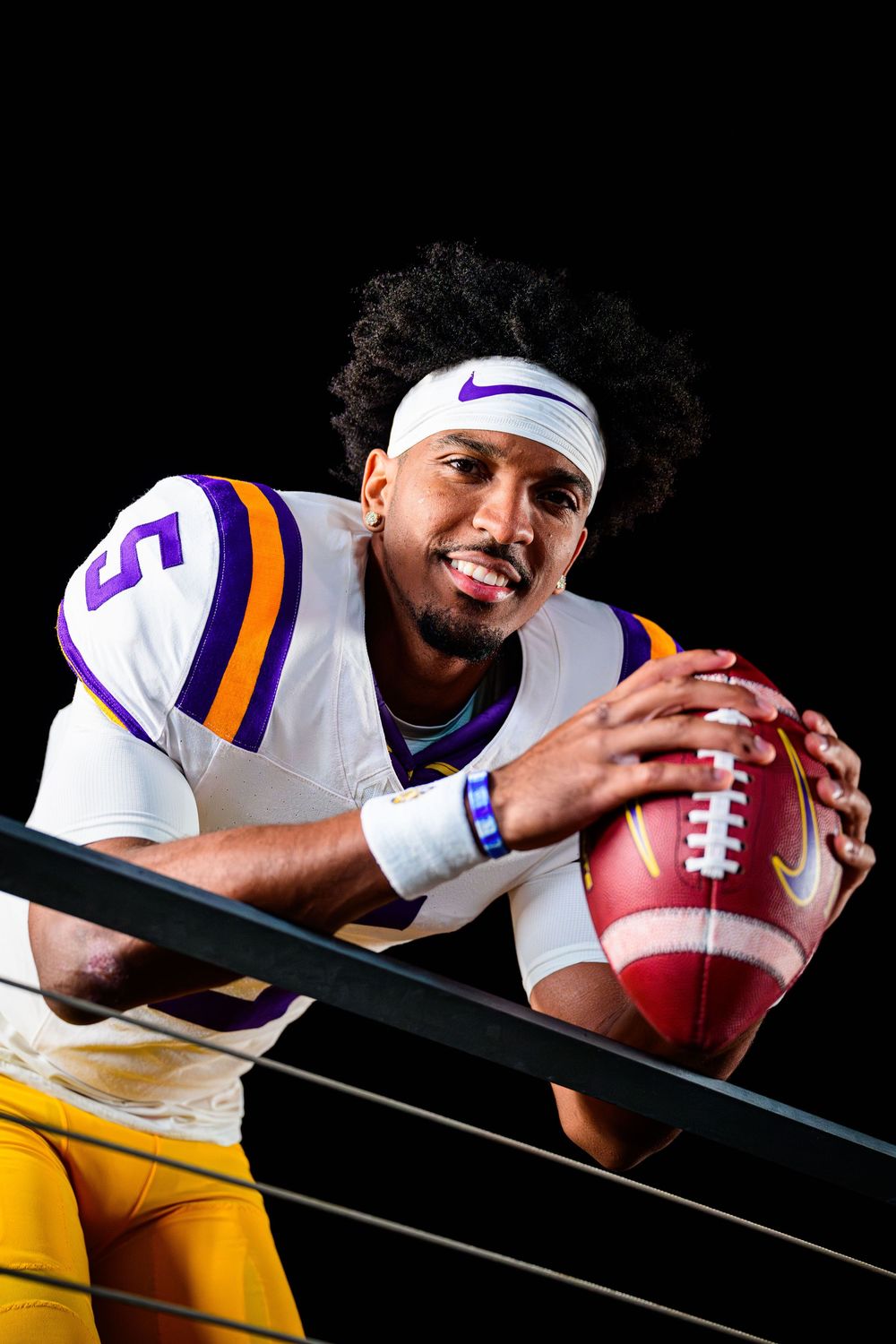 Football player in a white jersey with purple and gold trim and a white Nike headband smiles while leaning on a railing holding a football.