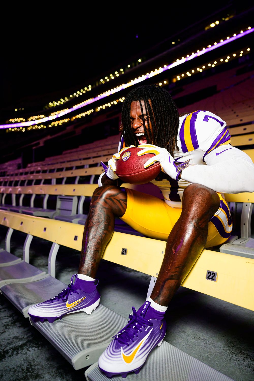 Football player in purple and gold uniform and cleats sits on empty stadium bleachers, gripping a football and yelling under glowing lights.