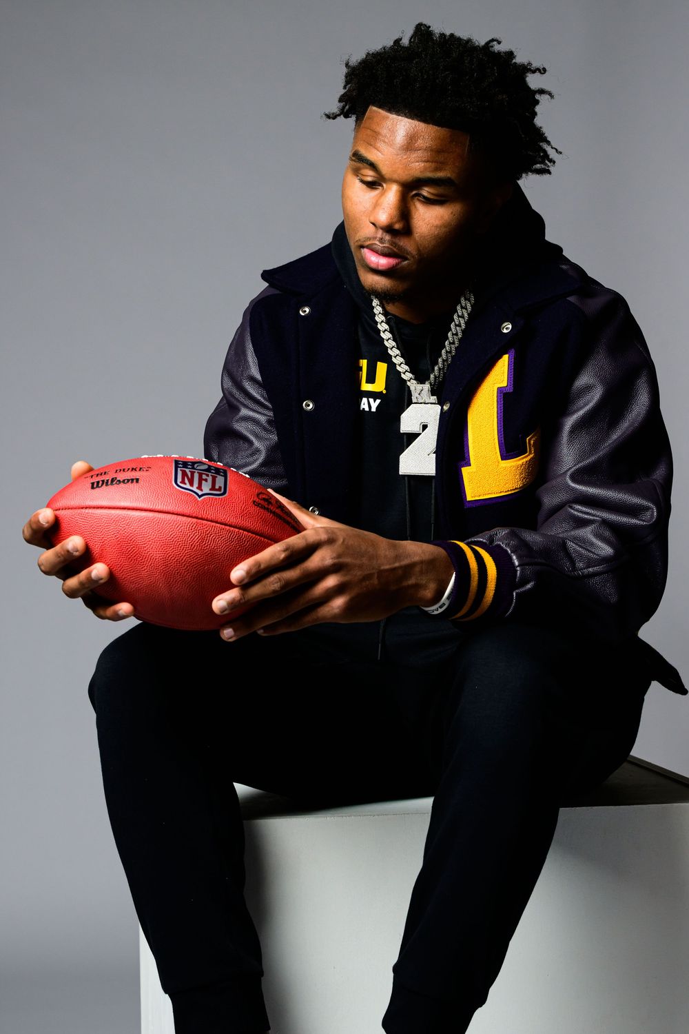 Young man in a navy and gold varsity jacket and chain necklace sits on a white block holding a Wilson NFL football against a gray backdrop.