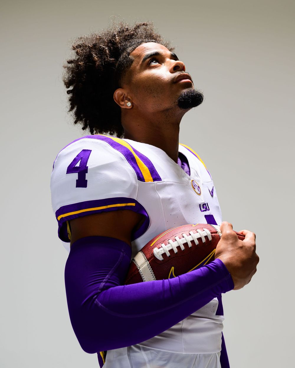 Football player in a white number 4 jersey with purple and gold trim and purple arm sleeve holds a football while looking upward.