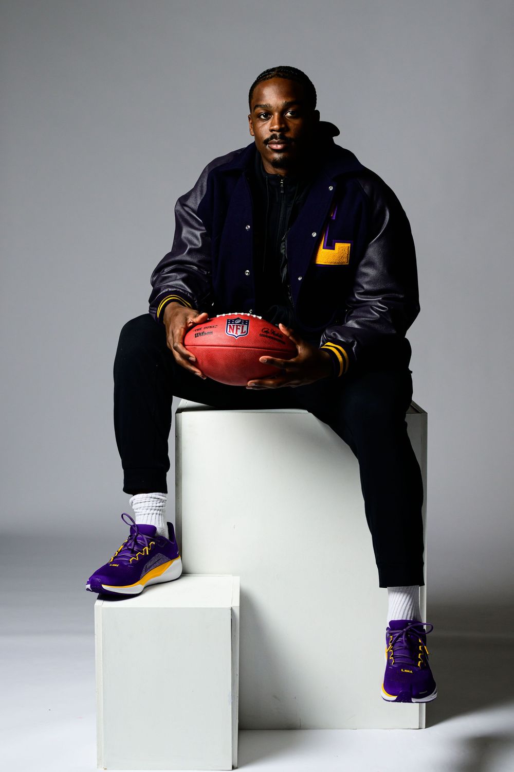 Professional athlete wearing varsity jacket and purple sneakers sits on white blocks holding NFL football in studio setting.