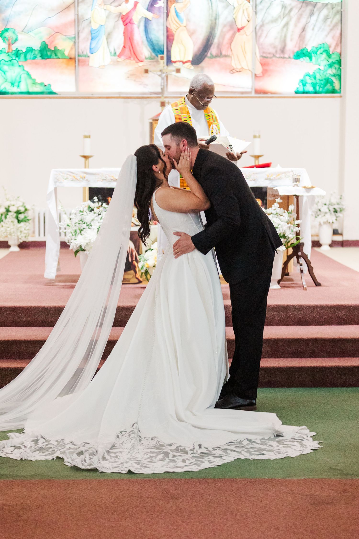 Bride in a long lace-trimmed gown and veil kisses the groom in a black suit before a priest at a church altar during their wedding ceremony.