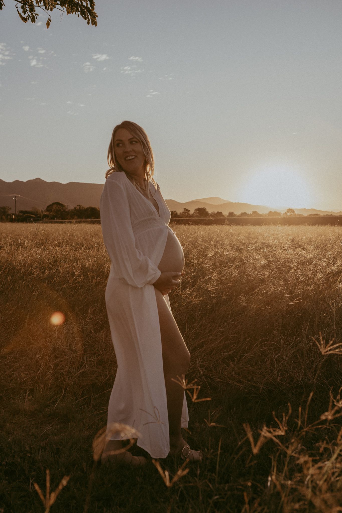 A glowing pregnant woman in a white dress smiles in a golden wheat field at stunning sunset.