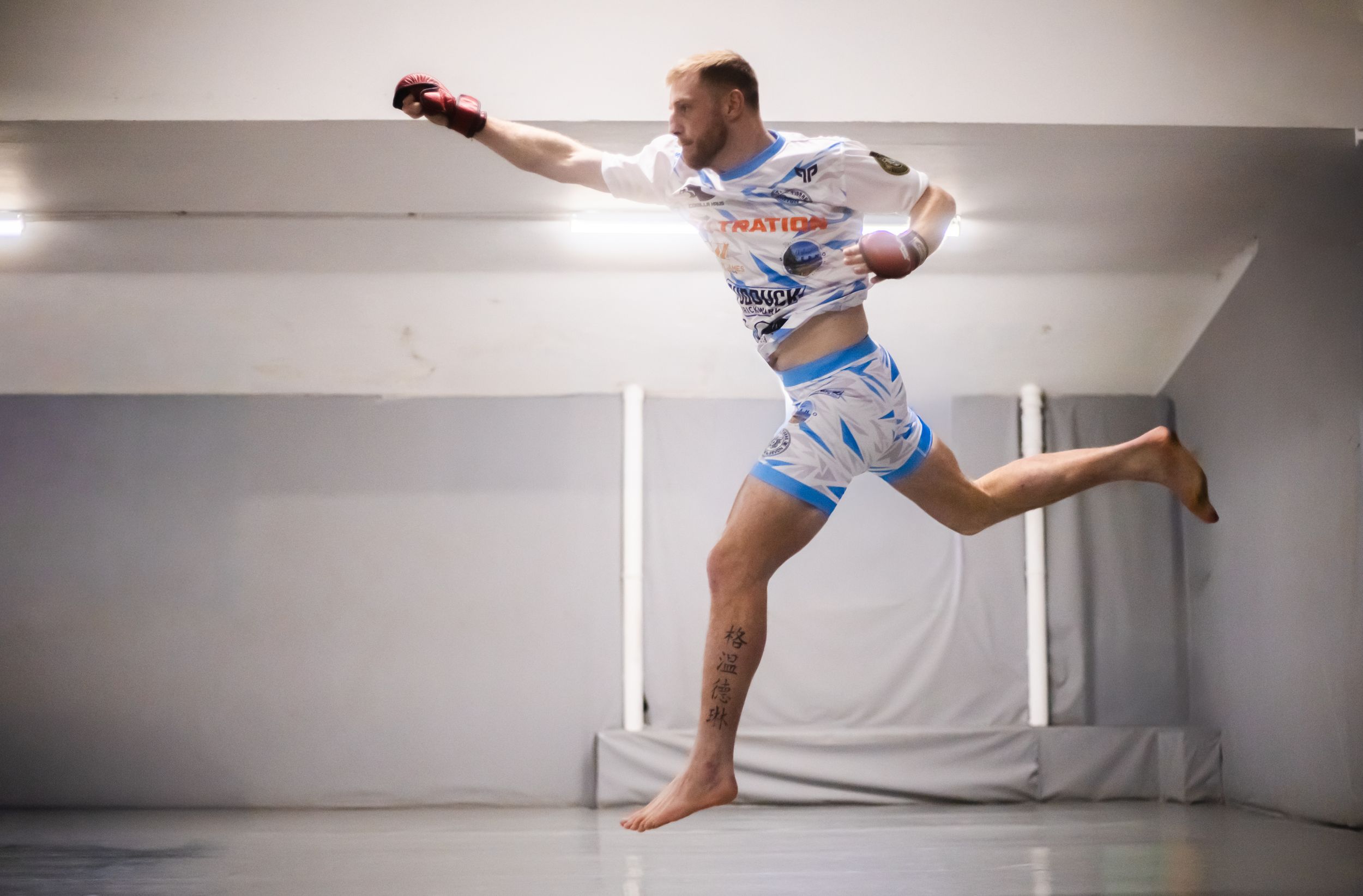 MMA fighter in white and blue shorts leaping mid-air with a punch during a training session indoors.