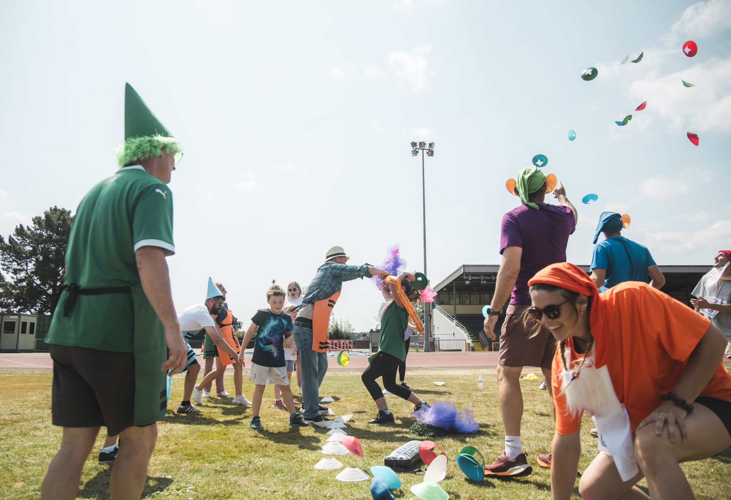 Children and adults in colorful costumes play outdoor games on a sunny field, tossing balloons during a festive event.