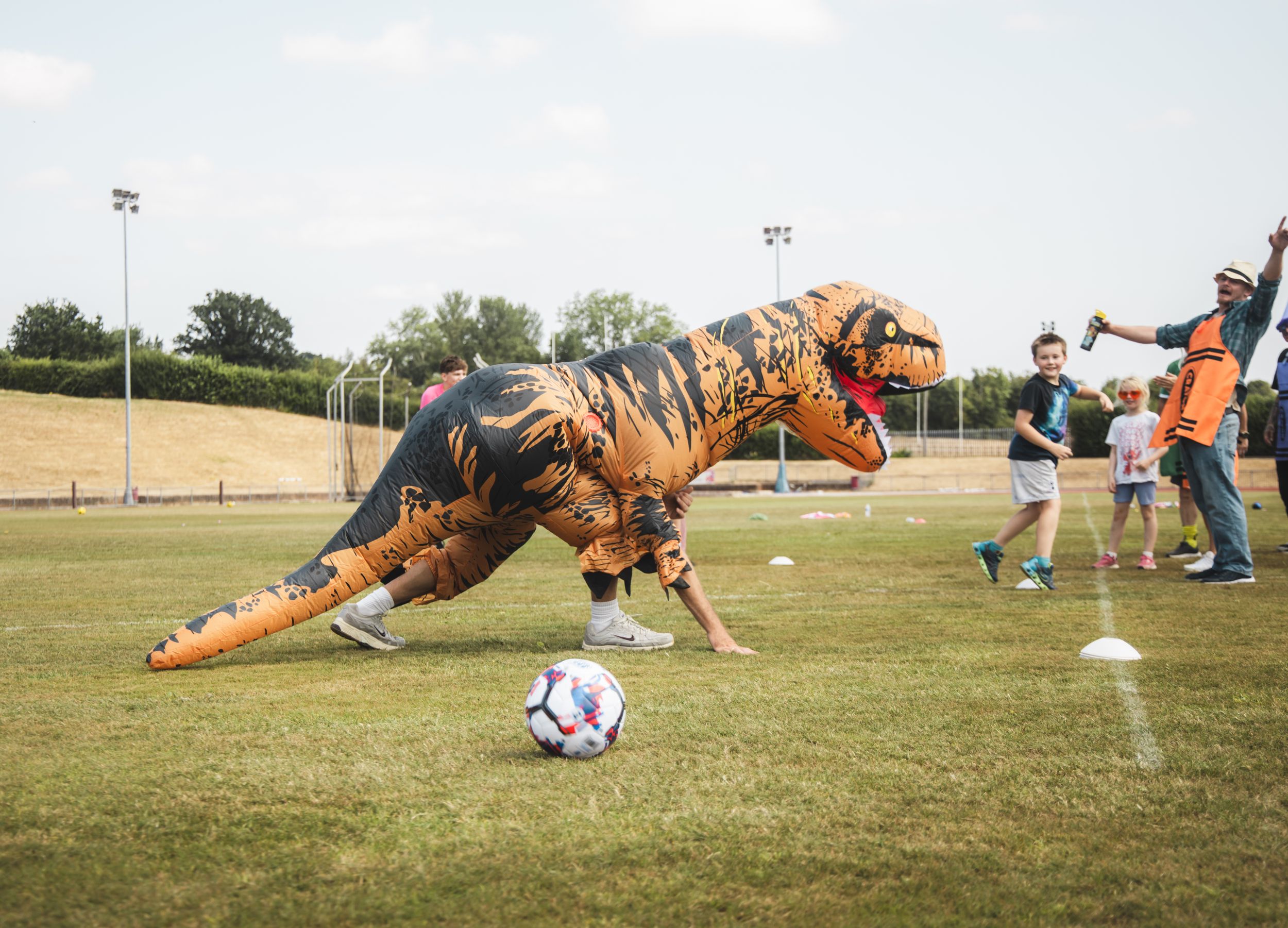 Person in inflatable T-Rex costume playing soccer on a green field with children during a fun outdoor event.