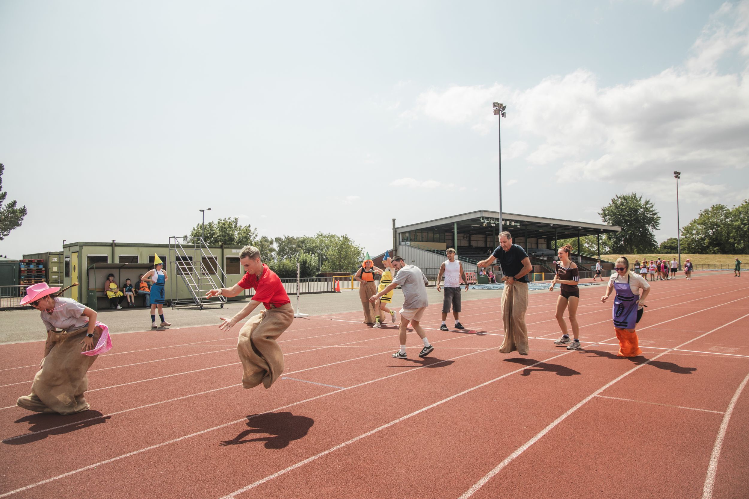 Adults competing in sack races on a red running track during a sunny outdoor sports day event.