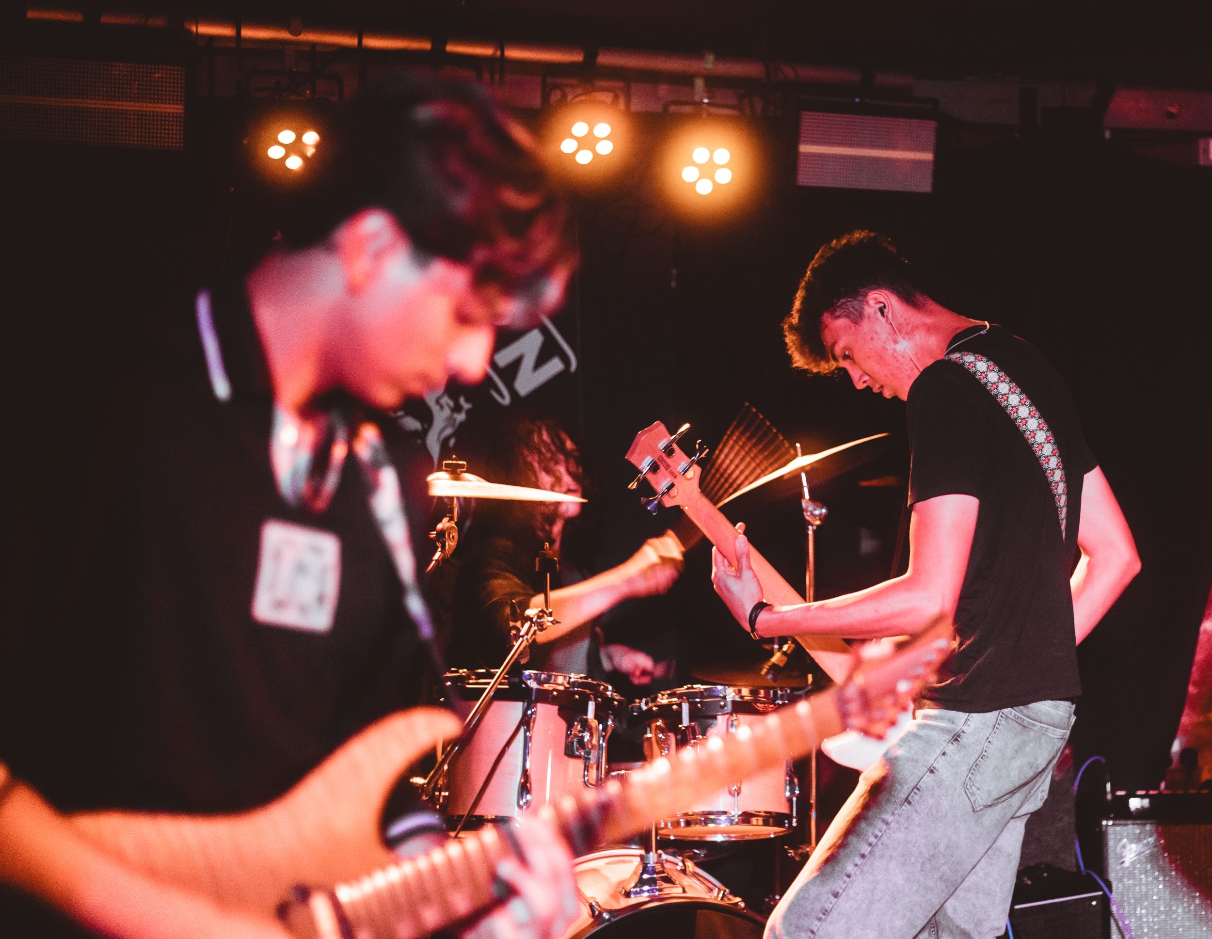 Young band performing live on stage with guitars and drums under warm orange stage lights in a dark venue.