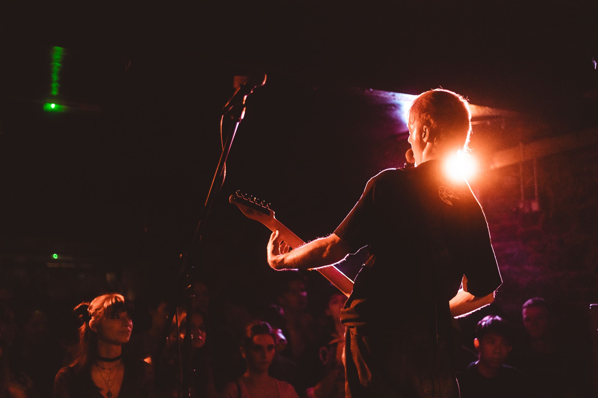 A guitarist performs on stage under warm orange lighting before an engaged crowd in a dark concert venue.