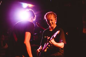 Two musicians performing on stage under purple lights, one playing electric guitar at a live concert.