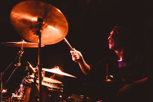 A drummer plays cymbals with drumsticks on a dark stage, illuminated by warm orange stage lighting.