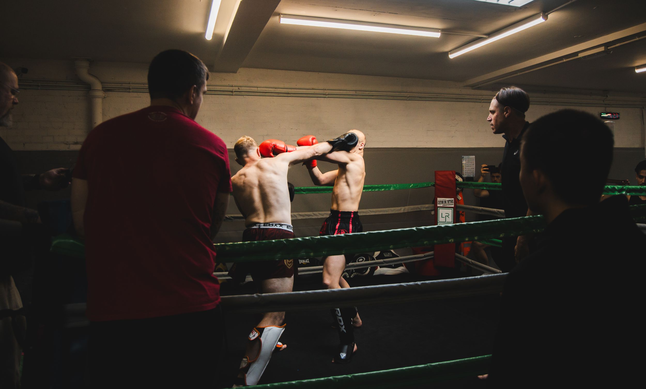 Two boxers sparring in a dimly lit boxing ring, watched by cornermen during an indoor boxing match.
