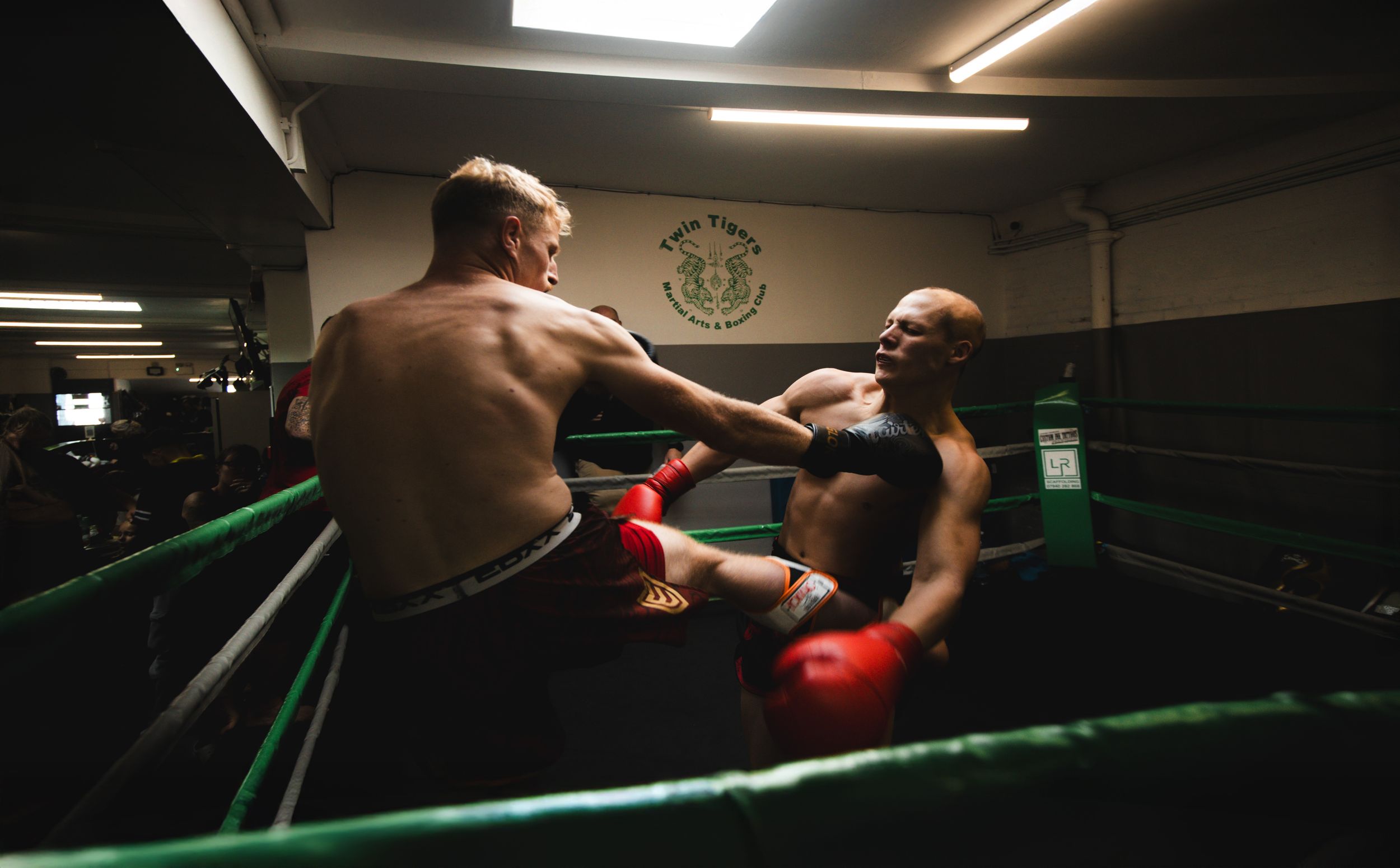 Two boxers sparring in a dimly lit boxing ring, wearing red gloves during an intense training match.