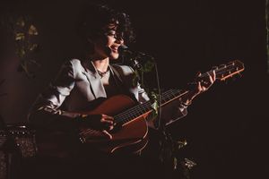 A musician with curly hair plays acoustic guitar and sings into a microphone on a dimly lit stage.