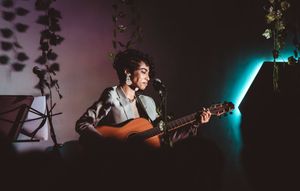 Young musician playing acoustic guitar and singing into a microphone on a dimly lit stage with plants.