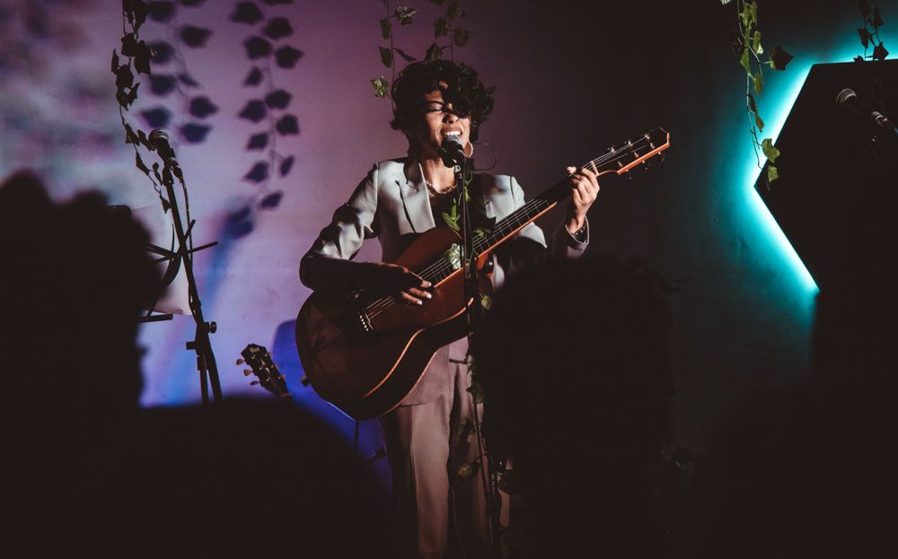 Musician playing acoustic guitar on stage with moody purple lighting and decorative foliage backdrop.