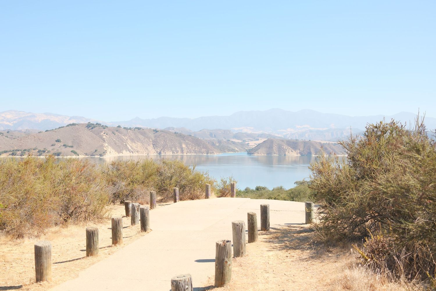 A scenic dirt pathway with wooden post barriers leading to a reservoir surrounded by dry hills and mountains under a clear blue sky.
