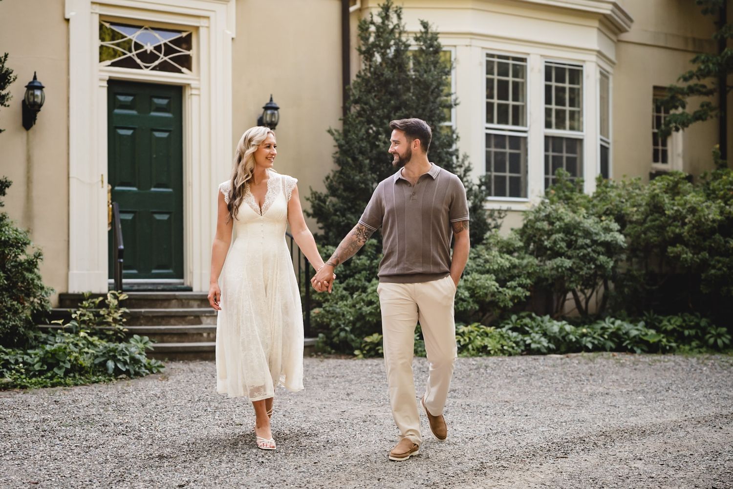 Couple holding hands walking on gravel path outside elegant yellow home with green door.