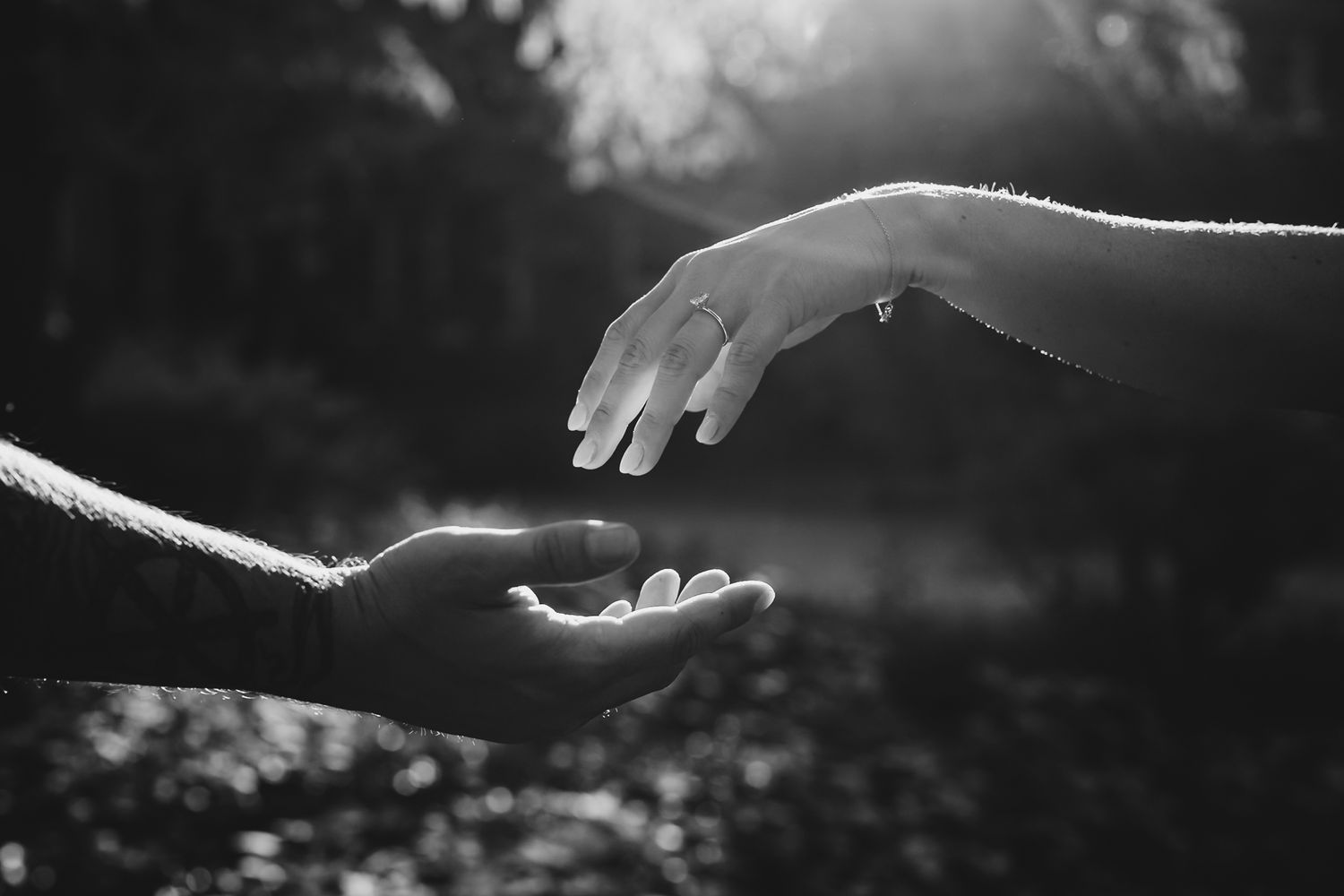 Black and white photo of two hands almost touching, one wearing a ring, with soft bokeh background lighting.