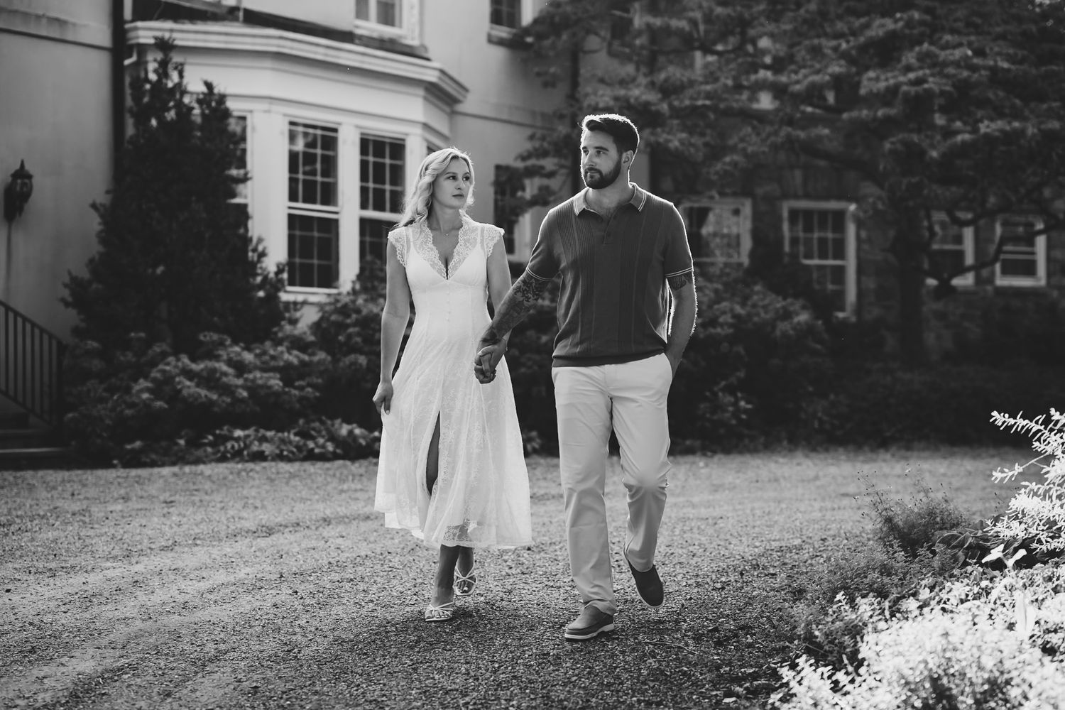 Black and white photo of a couple walking hand in hand on a garden path outside an elegant estate.