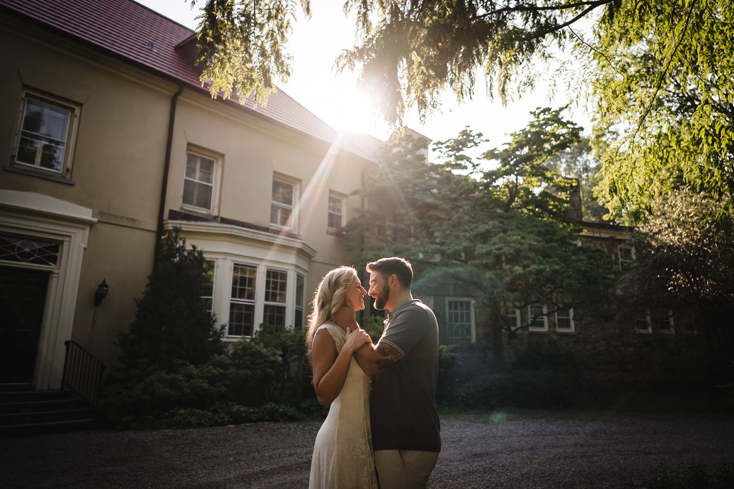 Couple sharing a romantic moment at golden hour outside an elegant manor house surrounded by lush greenery.