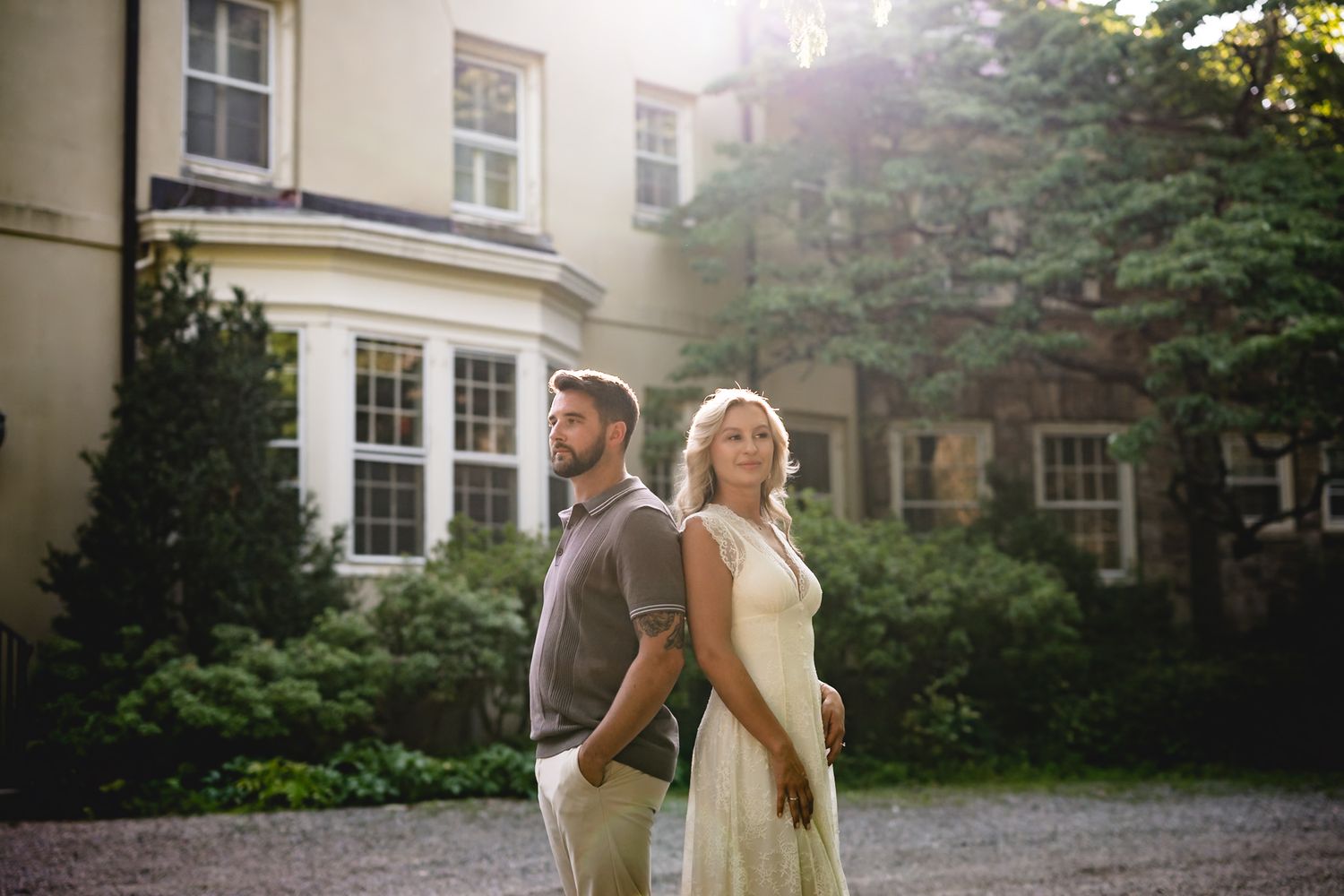 A couple stands back-to-back in a sunlit garden beside an elegant manor house, the woman in a cream dress and man in casual attire.