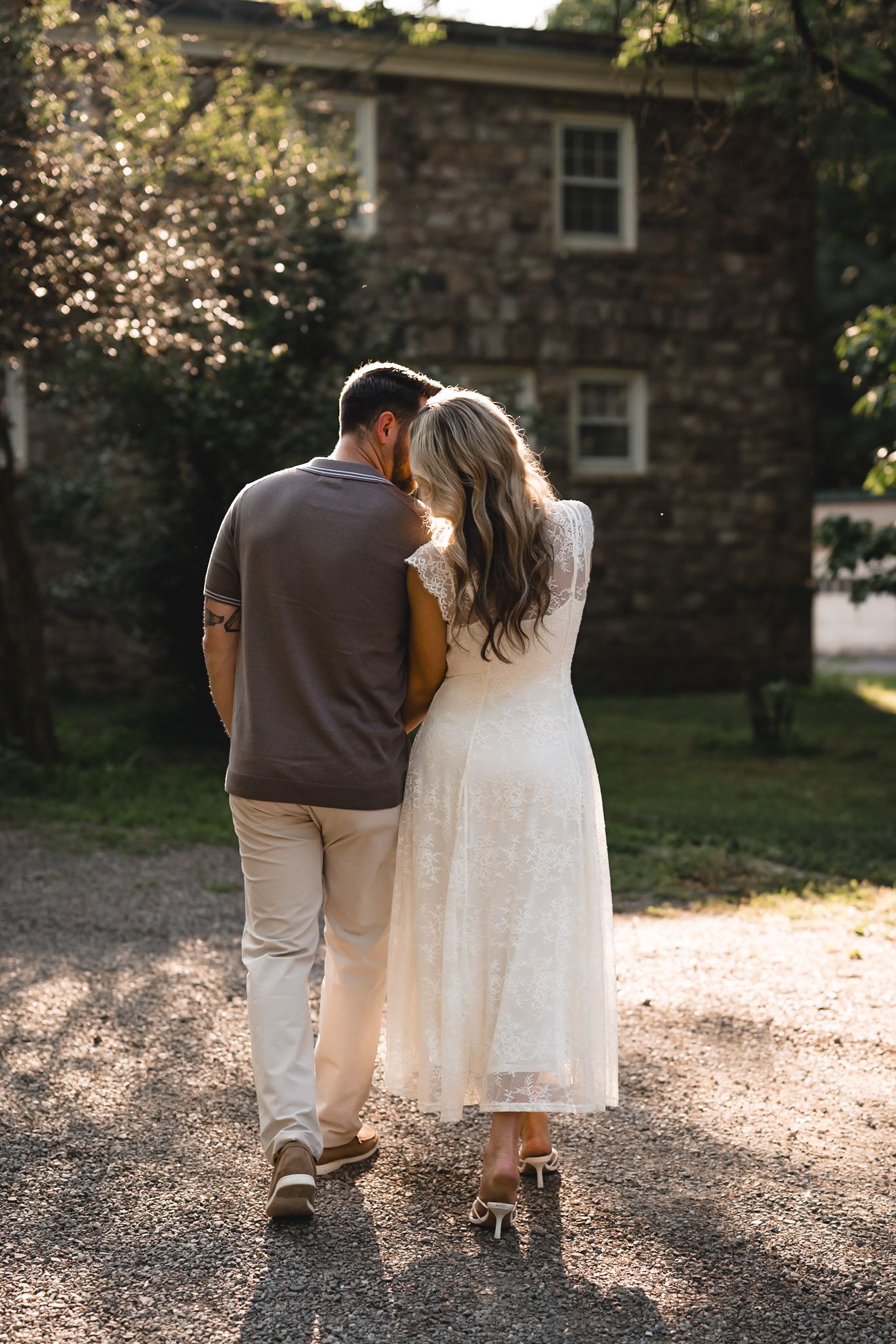 Couple walking hand in hand toward a stone house, woman in white lace dress and man in brown shirt, golden hour light.