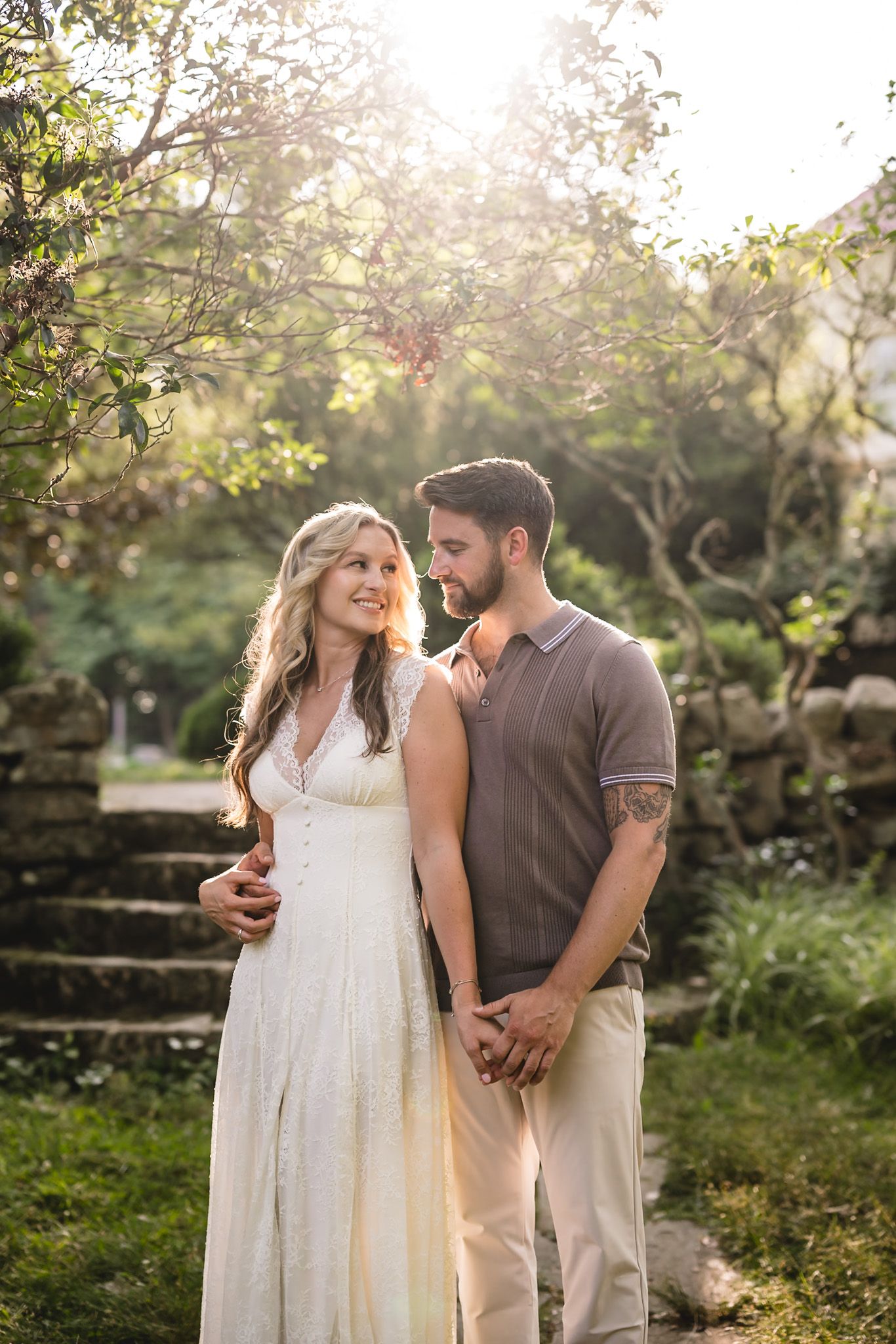 A couple in a sunlit garden, woman in white dress and man in brown shirt, holding hands and smiling at each other.