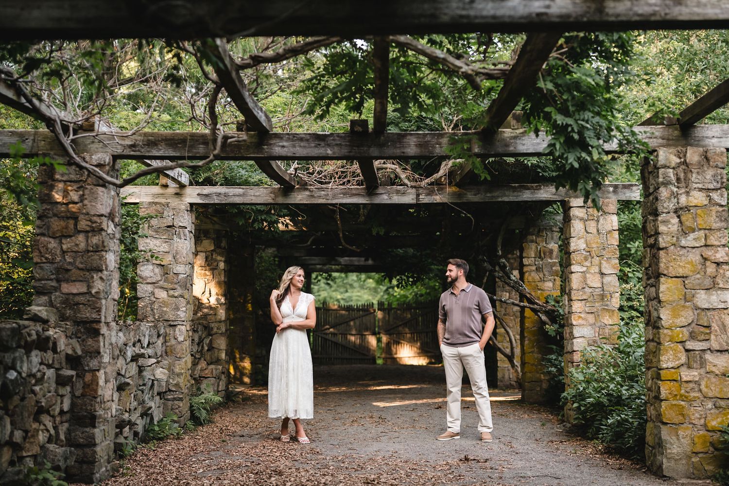 Couple standing apart under a rustic stone pergola covered in vines, surrounded by lush greenery in a garden setting.
