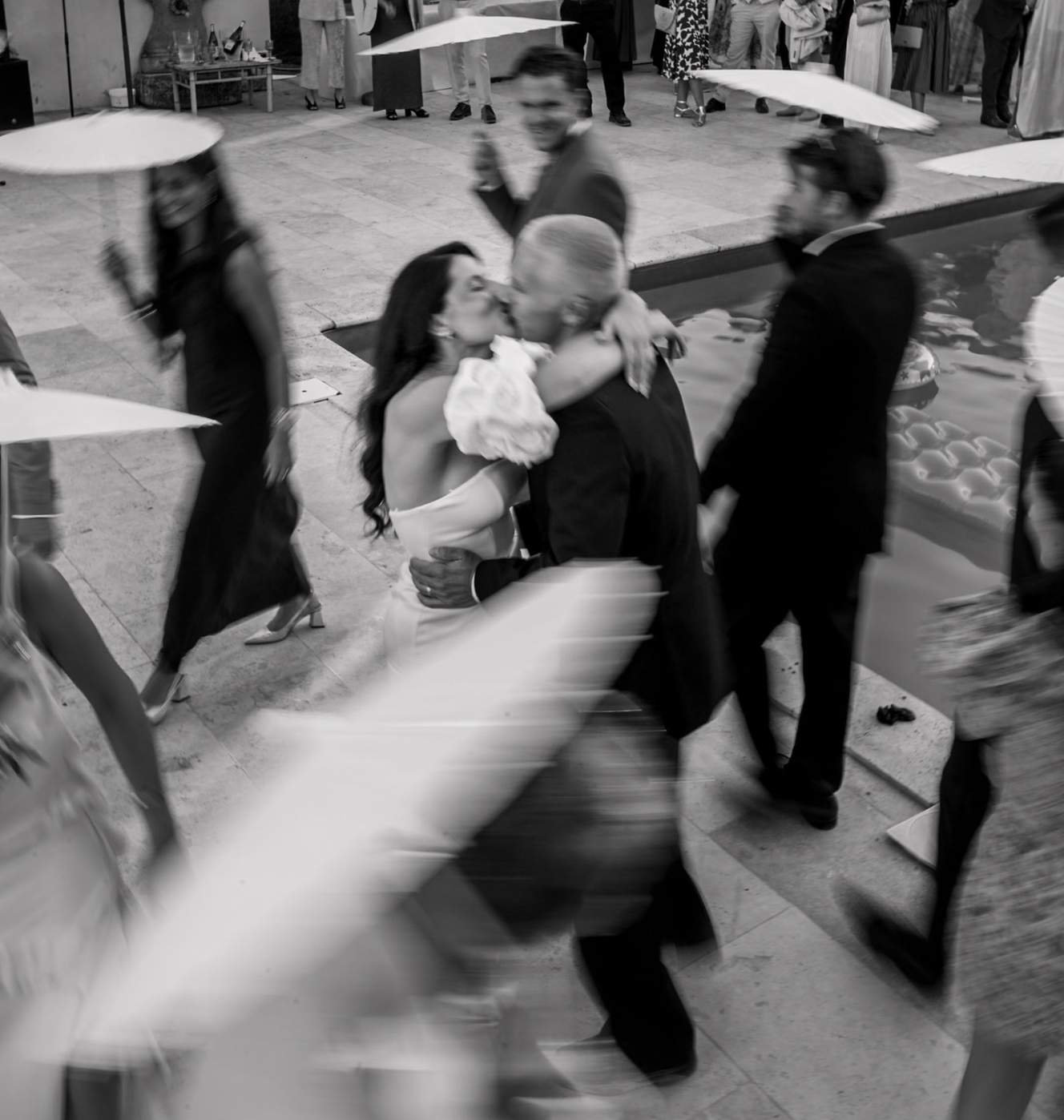 Black and white wedding photo of a bride and groom kissing near a pool while blurred guests in formal attire move around them.