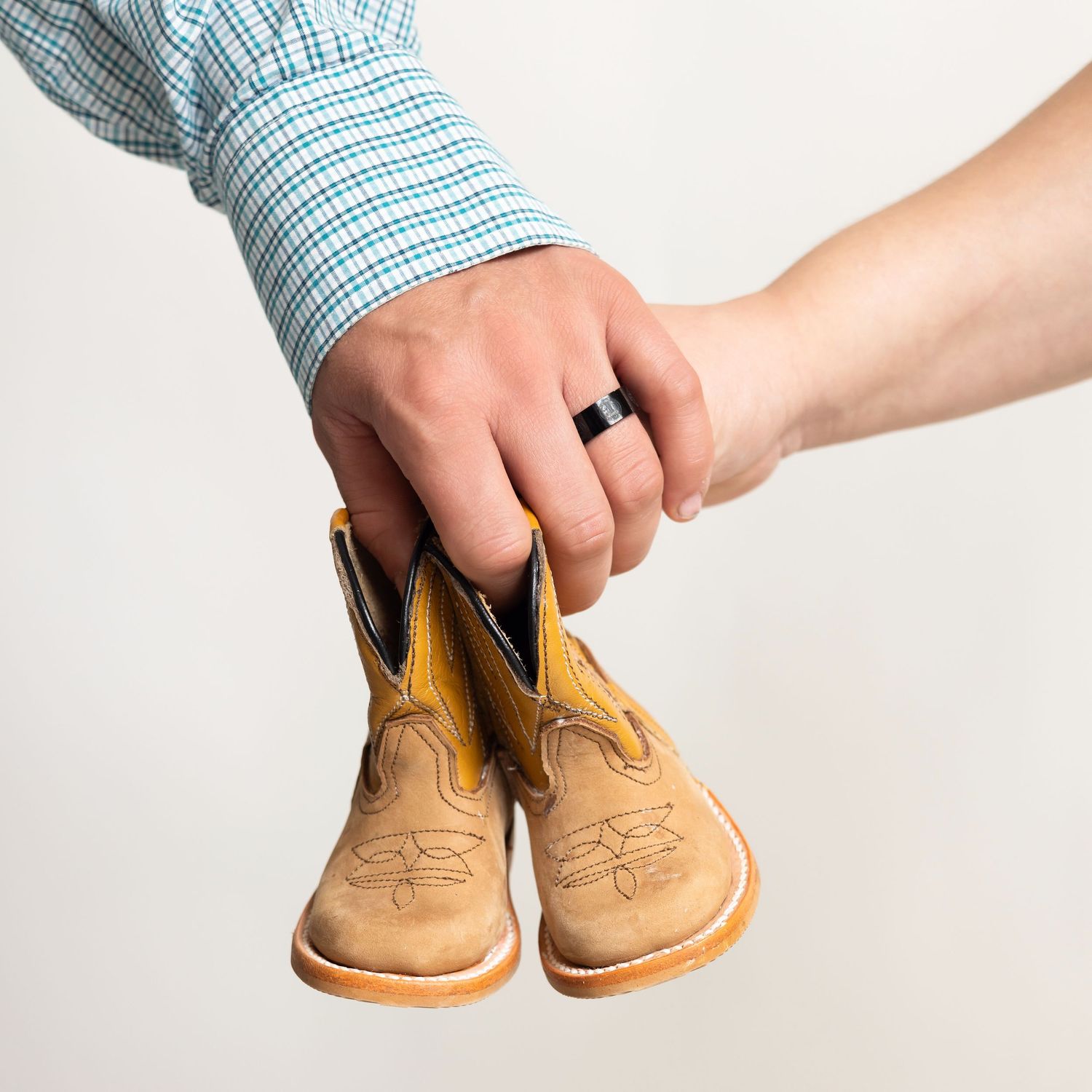 A man wearing a wedding ring holds tiny baby cowboy boots, suggesting a pregnancy announcement.